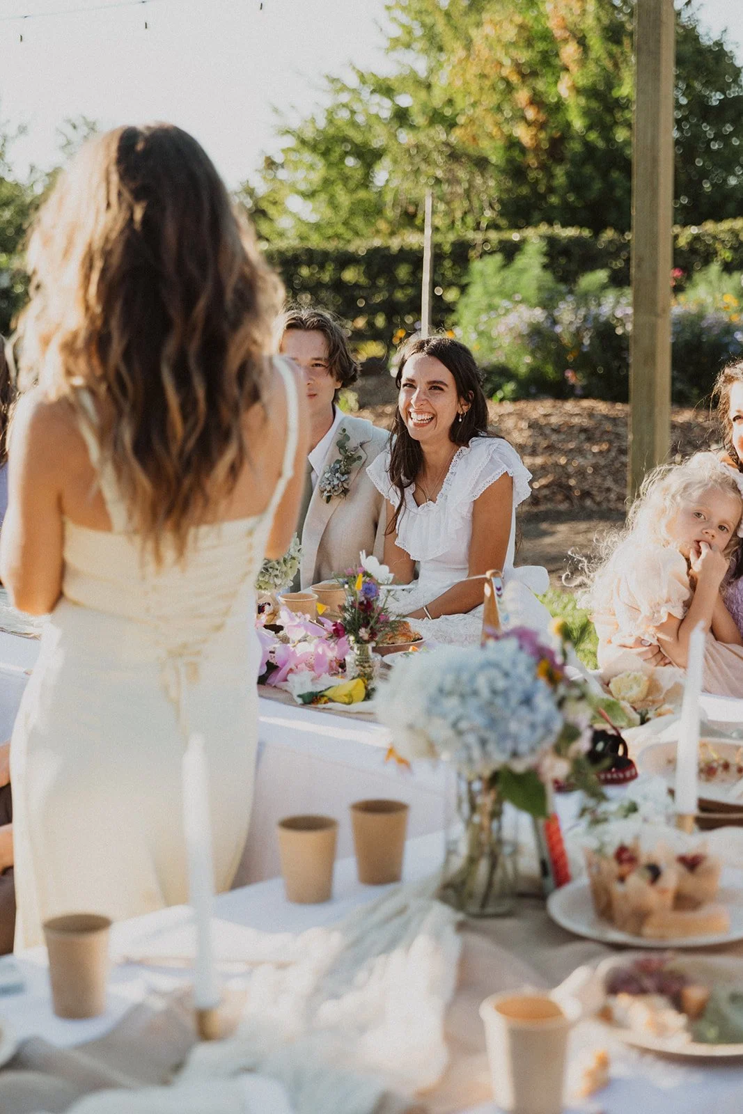 Bride smiling and reacting emotionally during a heartfelt speech at an outdoor wedding reception table with guests and florals