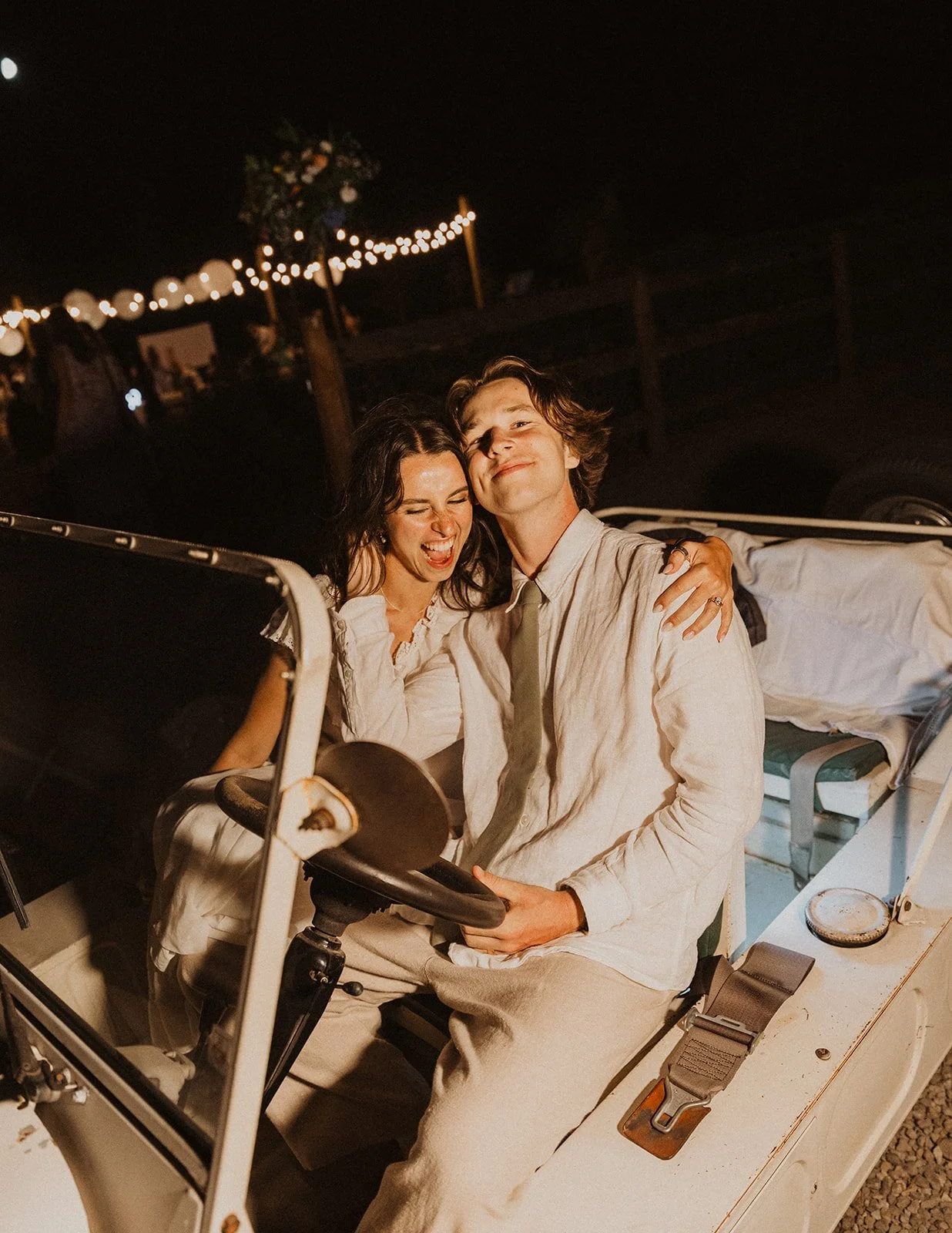 Newlywed couple smiling and sitting together in a vintage car during their wedding send-off with glowing string lights in the background