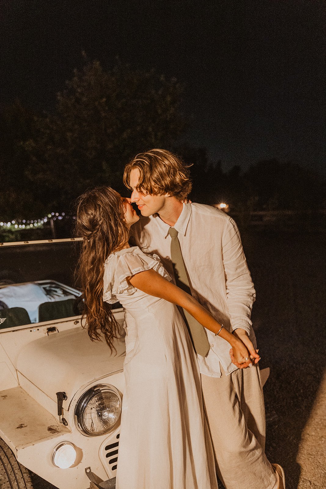 Bride and groom leaning in for a kiss beside a vintage car under warm string lights during a nighttime backyard wedding
