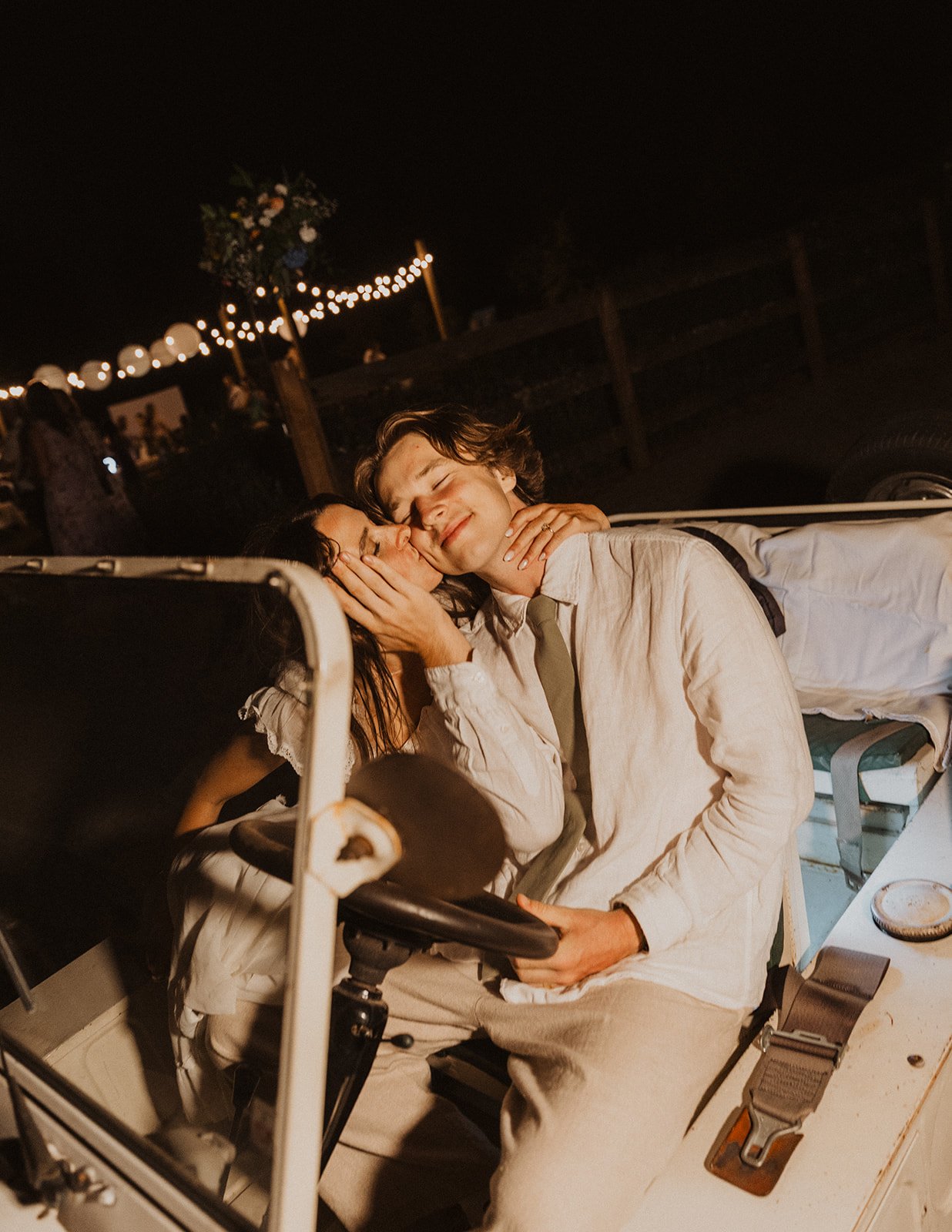 Bride kissing groom’s cheek as they sit in a vintage car during a romantic nighttime wedding exit with soft string lights behind them