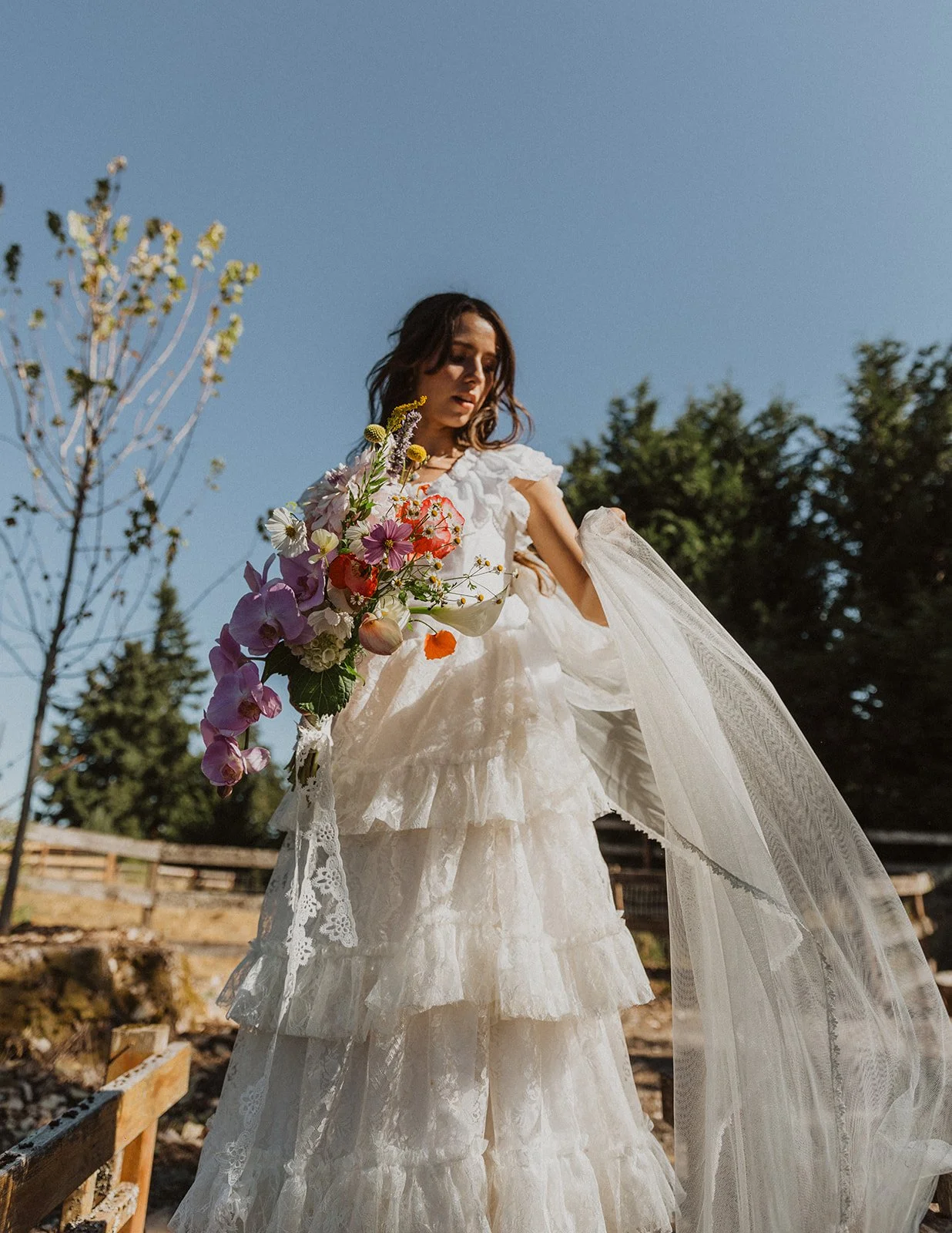 Bride in a layered lace wedding dress holding a colorful wildflower bouquet with flowing veil in a sunlit outdoor setting