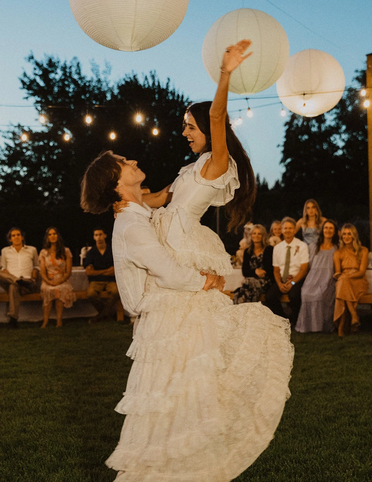 Groom walking behind bride holding her long veil as they cross a small wooden bridge in a lush garden wedding setting