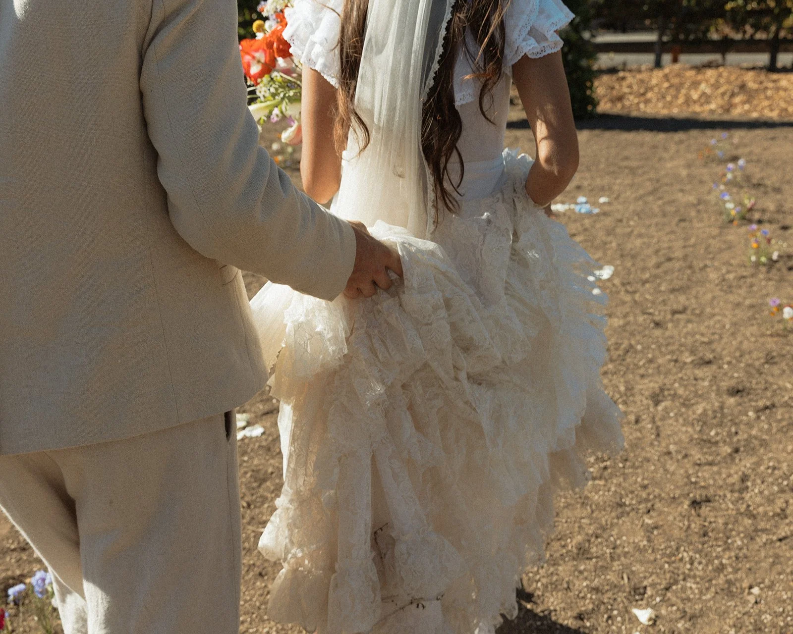 Groom gently holding the back of the bride’s lace wedding dress as they walk together down a sunlit outdoor path
