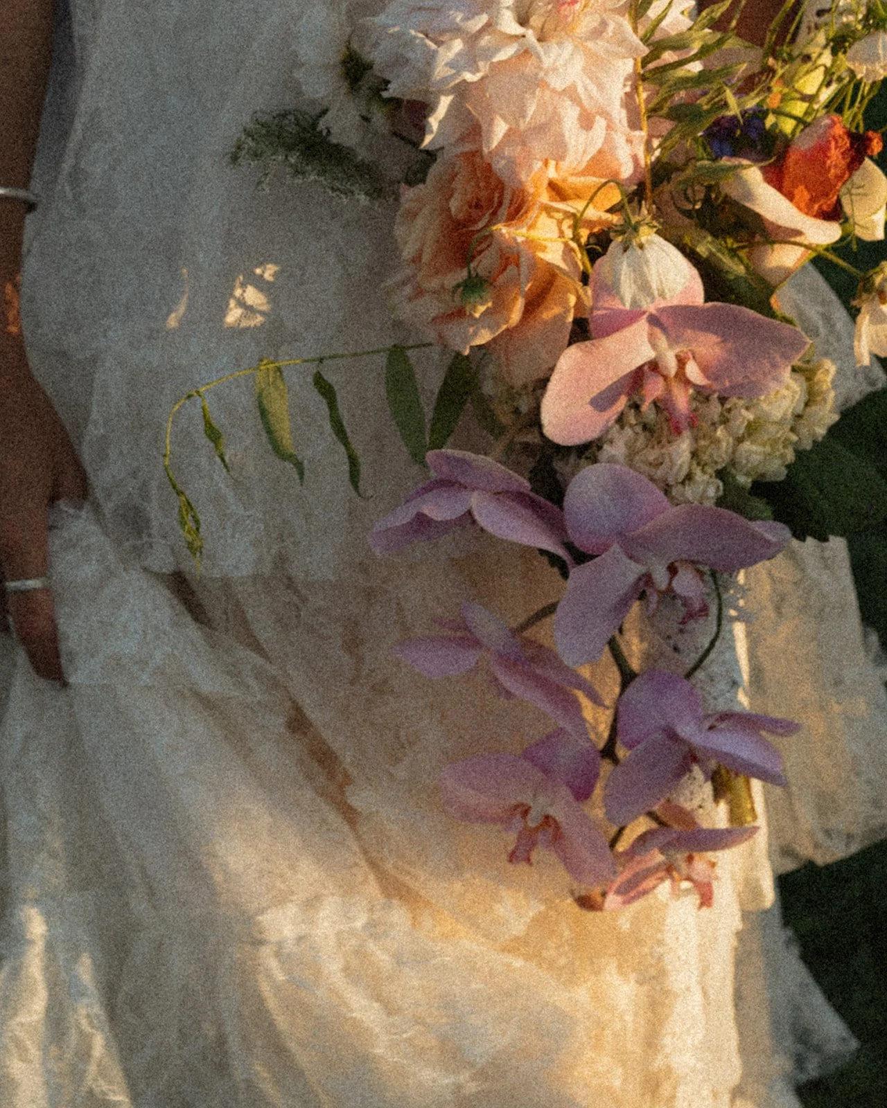 Close-up of bride’s hand holding a soft, romantic bouquet with orchids and pastel florals against a lace wedding dress