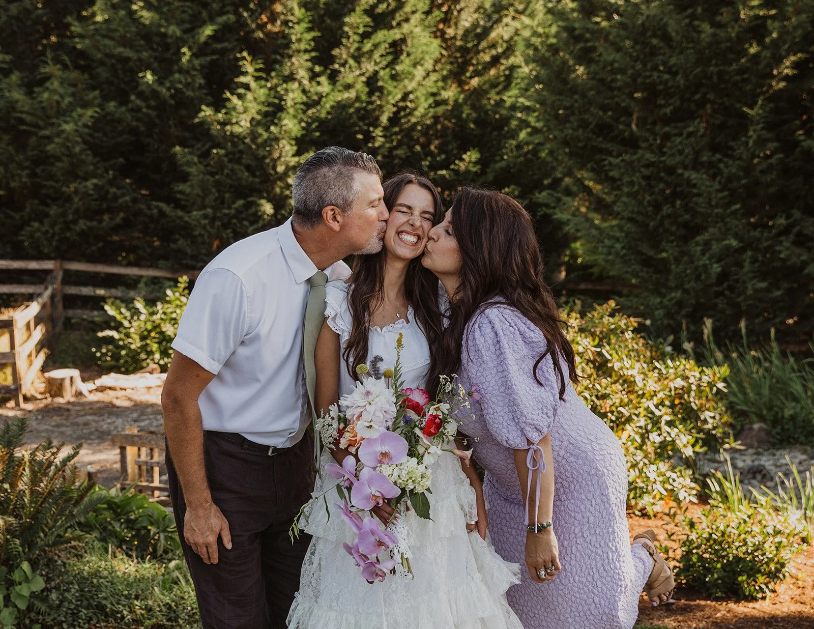 Bride smiling as her parents kiss her cheeks while she holds a colorful bouquet during an outdoor garden wedding moment