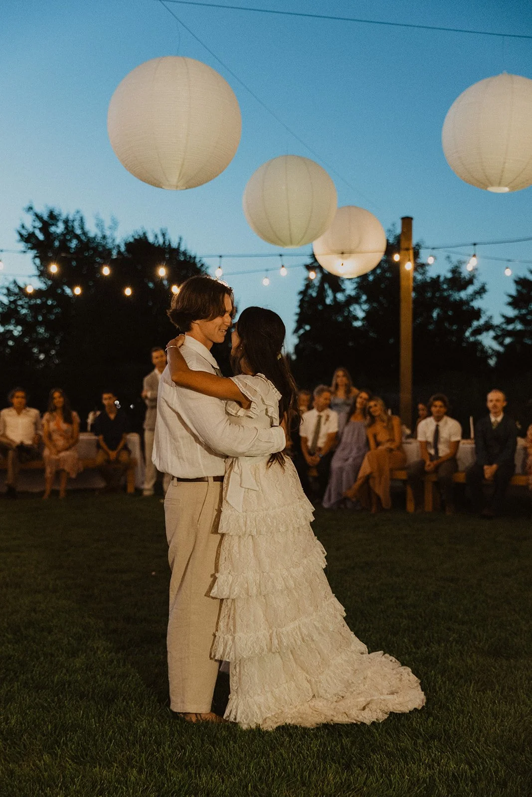 Bride and groom slow dancing under string lights and paper lanterns during an outdoor evening wedding, surrounded by seated guests