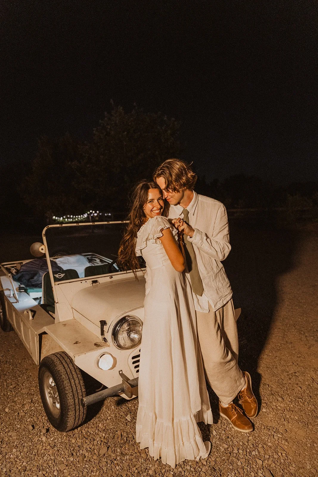 Bride and groom embracing beside a vintage car at night, sharing a quiet, intimate moment under soft ambient lighting