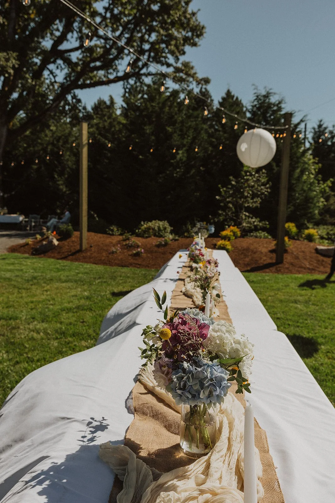 Long outdoor reception table styled with florals, candles, and linen runner set beneath string lights and paper lanterns in a backyard setting
