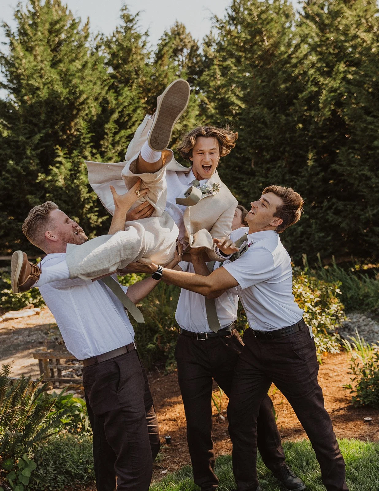 Groom laughing as he is lifted into the air by groomsmen in a playful moment during an outdoor wedding celebration surrounded by trees