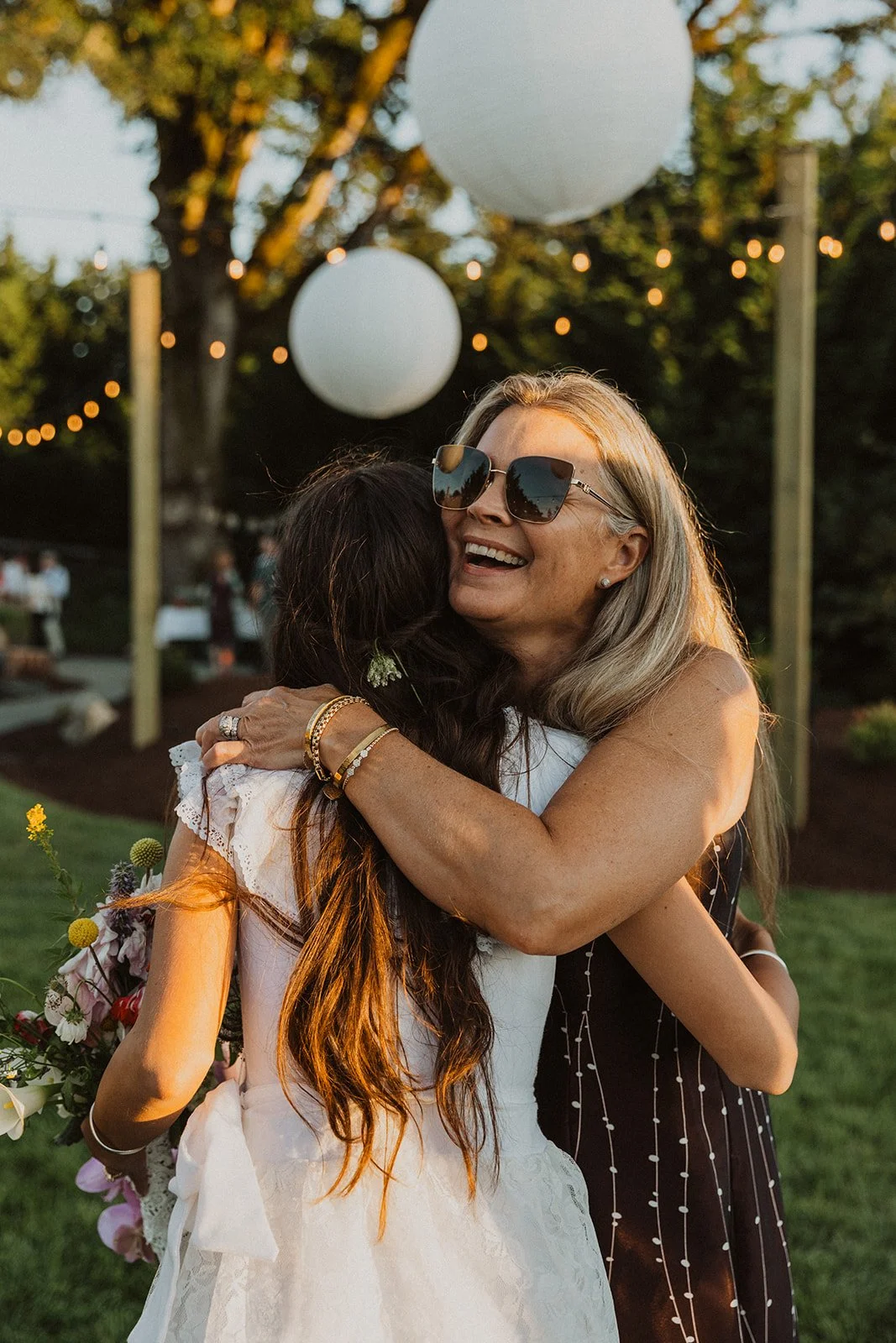Bride in a lace wedding dress hugging an older woman wearing sunglasses under string lights at an outdoor reception