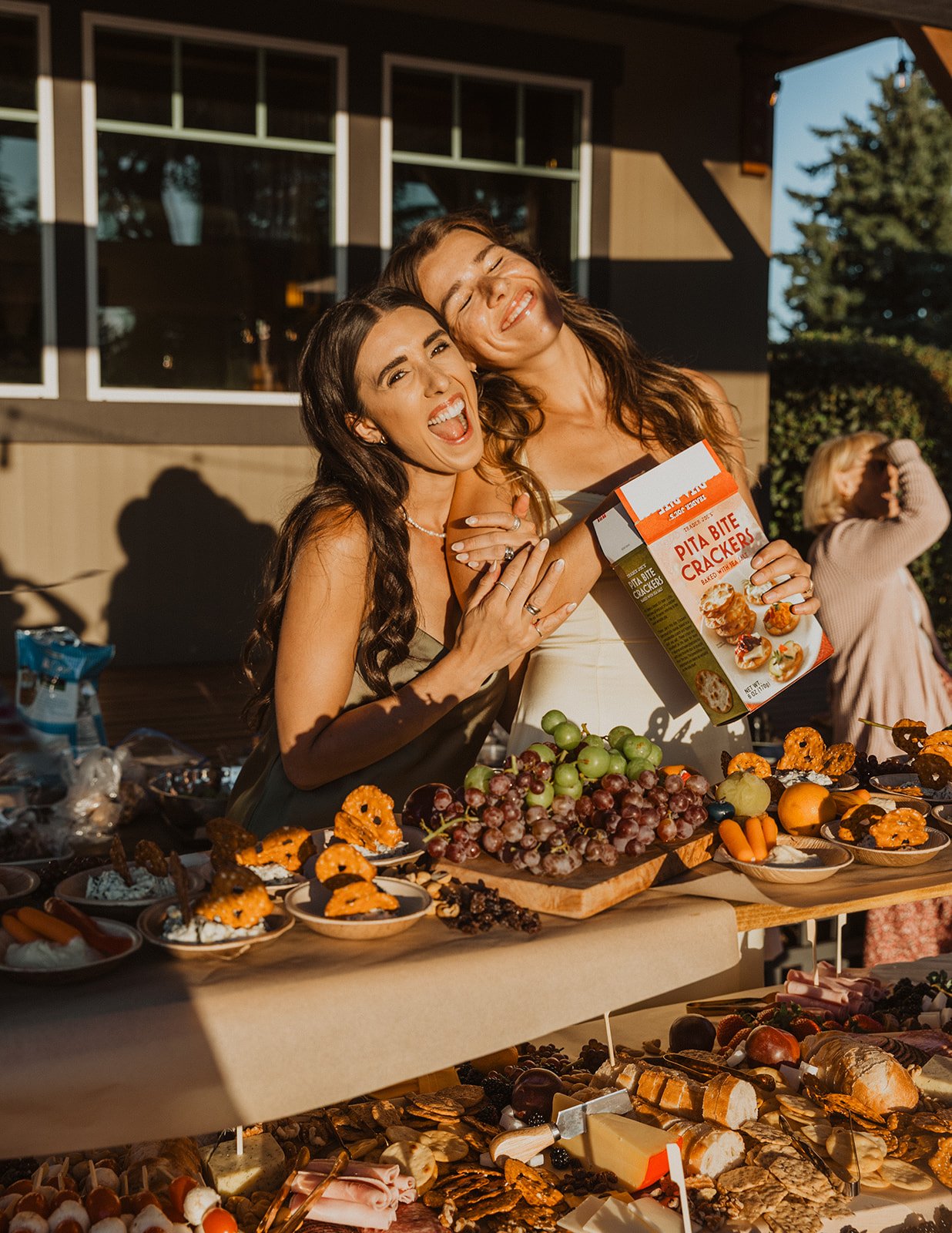 Two women laughing and posing behind a charcuterie-style food table with fruit, snacks, and appetizers at a wedding reception