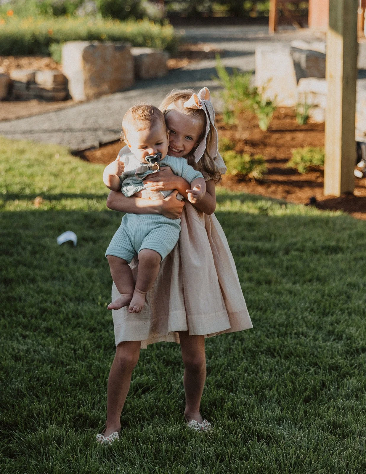 Young flower girl in a blush dress holding and hugging a baby on a grassy lawn during an outdoor wedding
