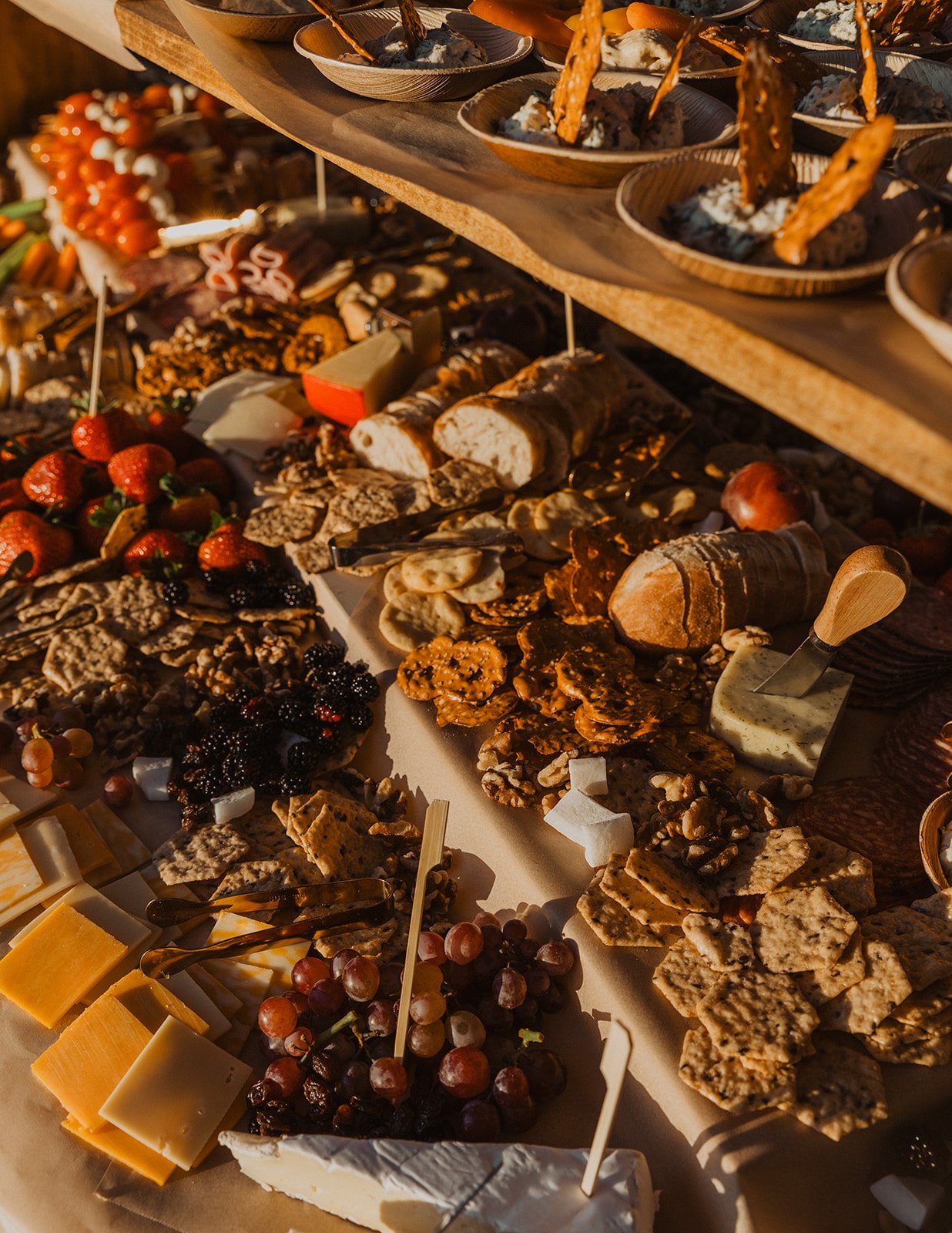 Close-up of a wedding grazing table with cheeses, meats, fruit, crackers, and appetizers in warm natural light