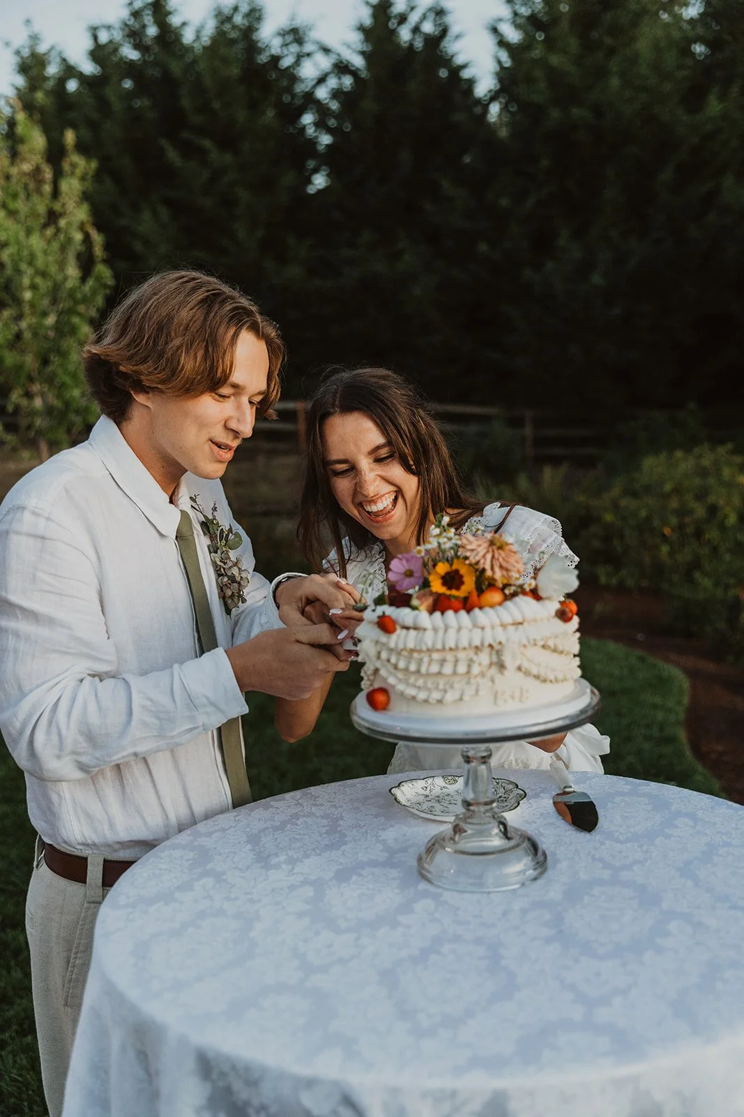 Bride and groom smiling beside a decorated wedding cake topped with fresh flowers and fruit during an outdoor reception