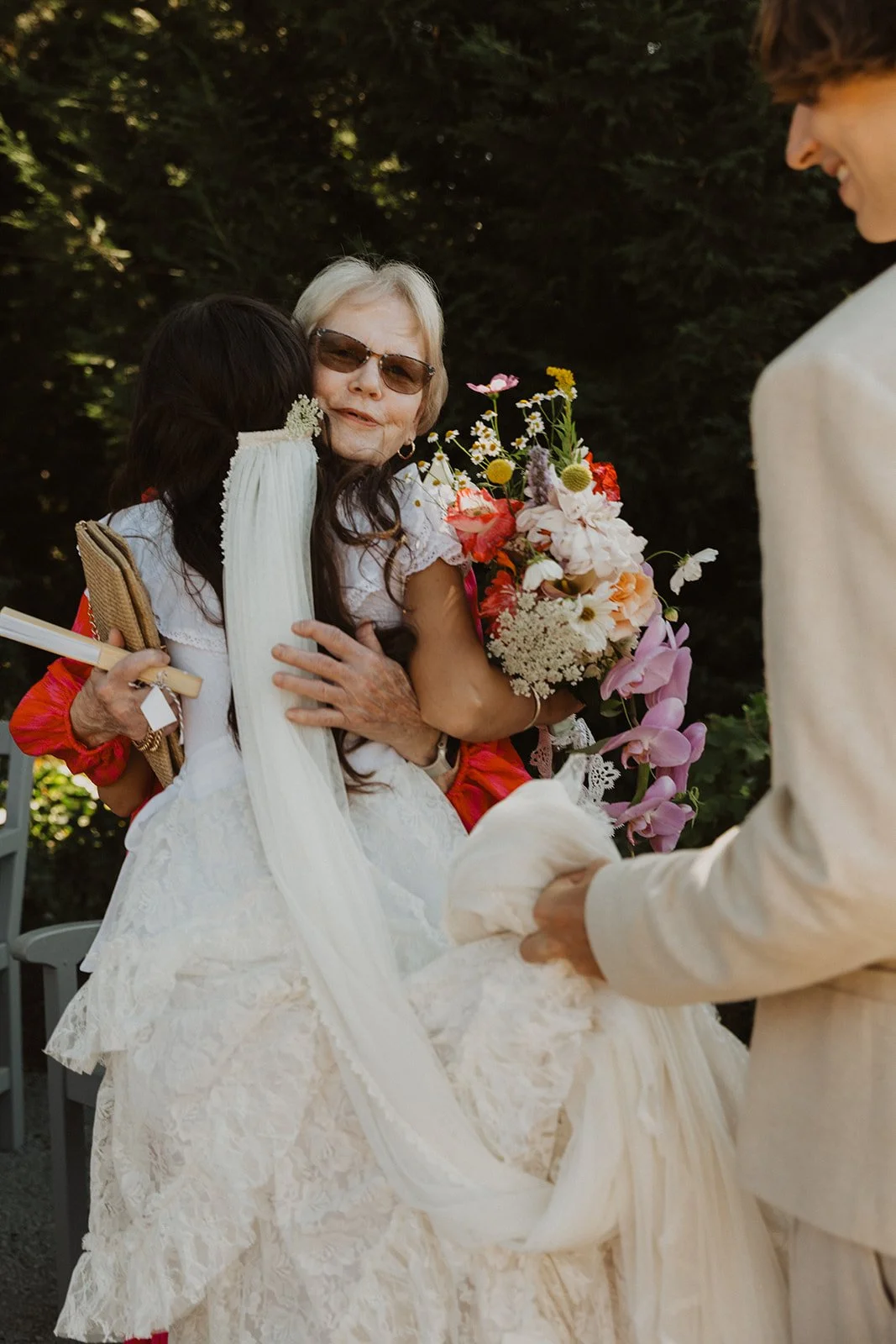 Bride in a lace wedding dress hugging an older woman while holding a colorful bouquet, with the groom nearby during an outdoor ceremony