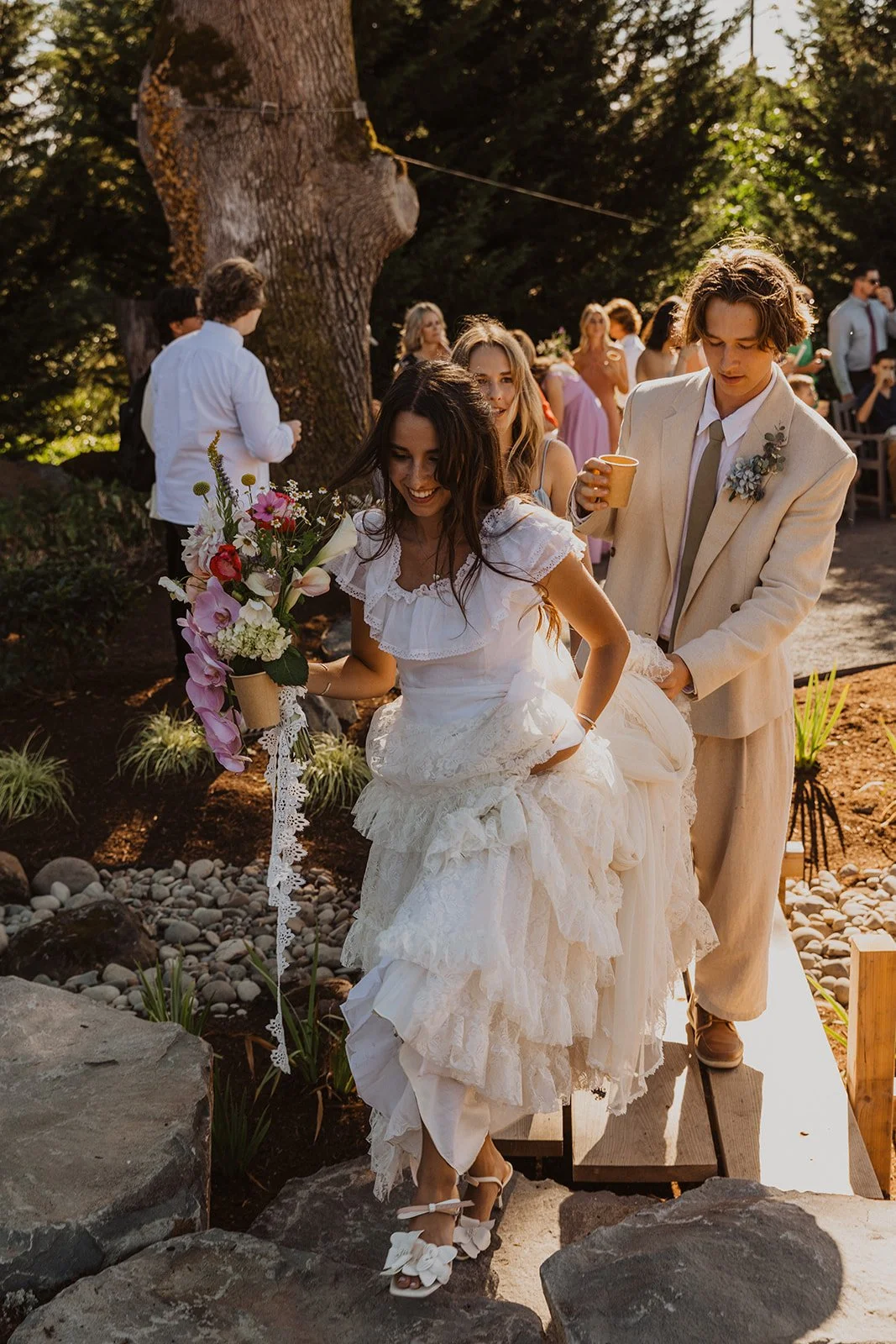 Close-up of the bride’s layered lace wedding dress and veil trailing across green grass in soft sunlight