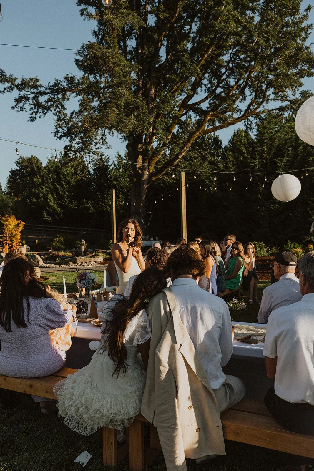 Guest giving a speech with a microphone during an outdoor wedding reception, while the couple and guests sit at a long table under string lights and a large tree