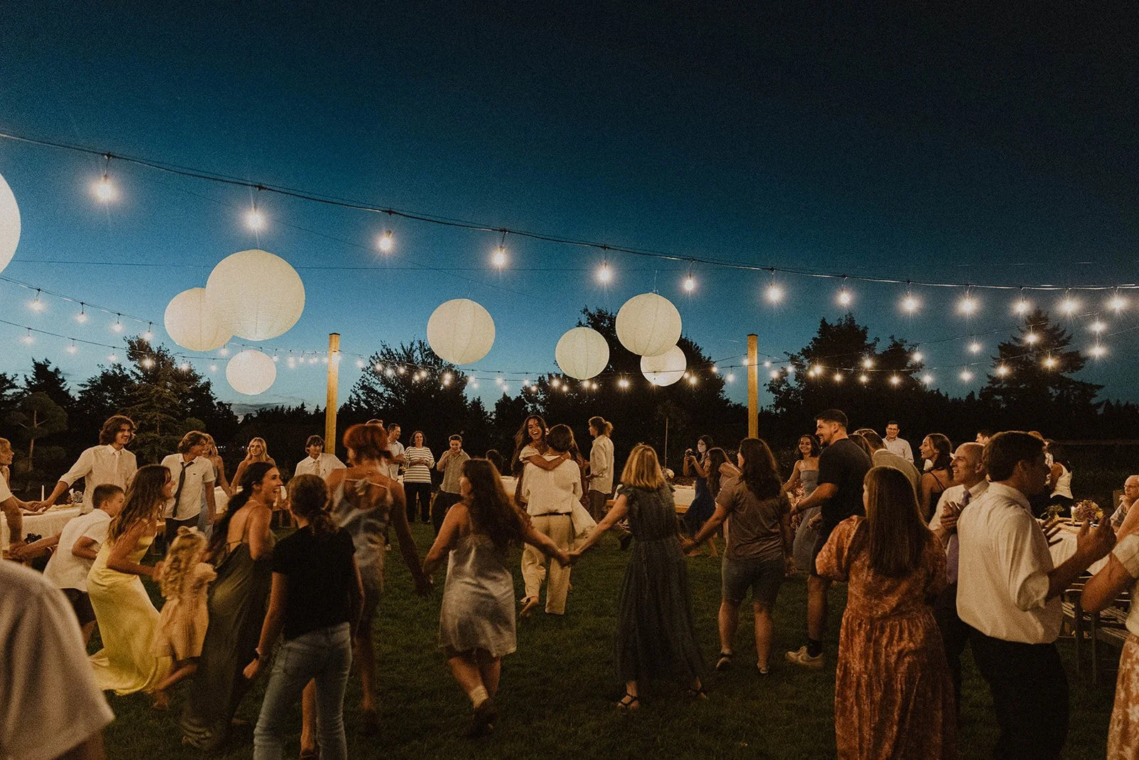 Guests dancing in a circle on a lawn at an outdoor wedding reception at night, with string lights and large paper lanterns glowing overhead