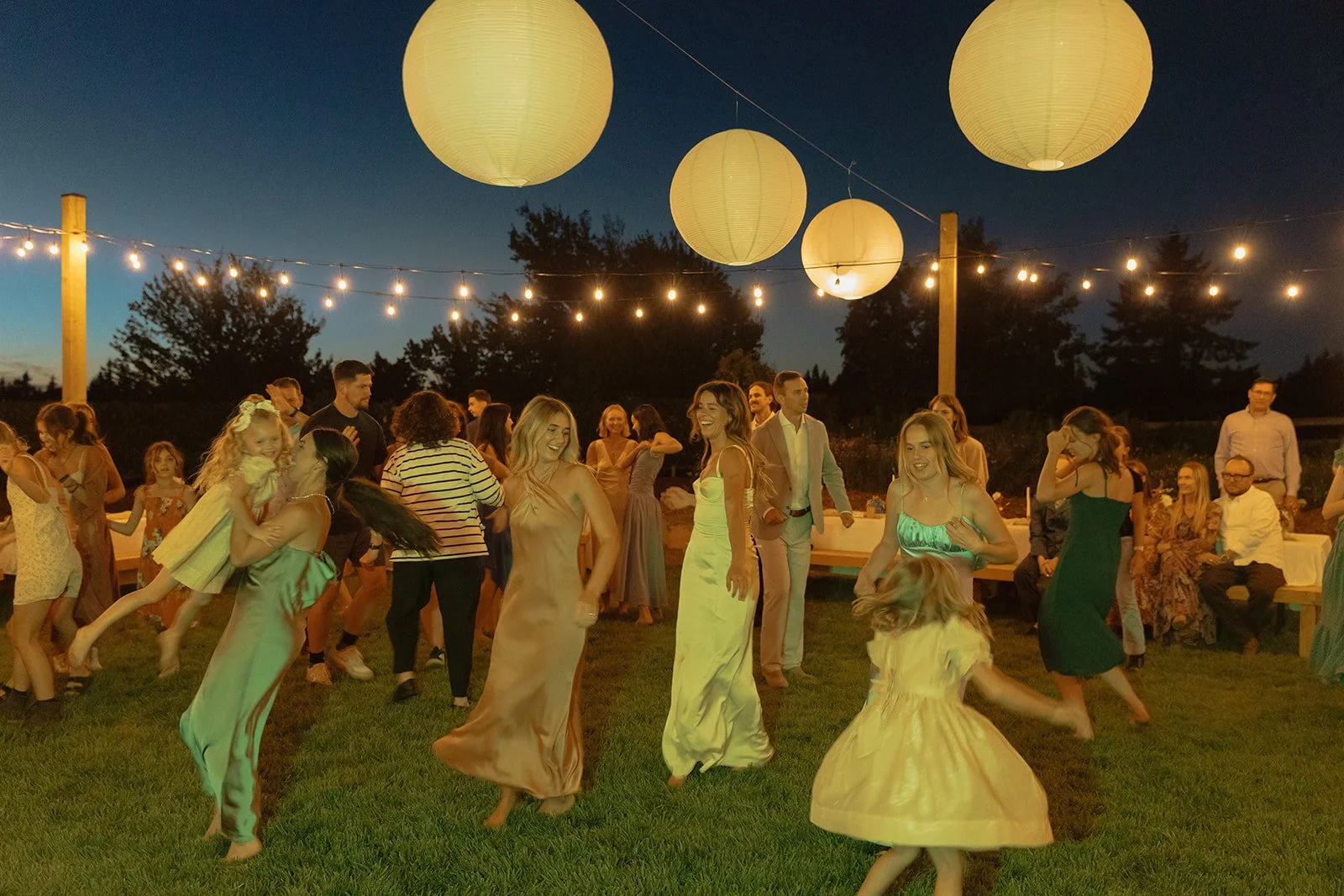 Wedding guests dancing and celebrating on the grass under string lights and hanging lanterns during a lively nighttime reception
