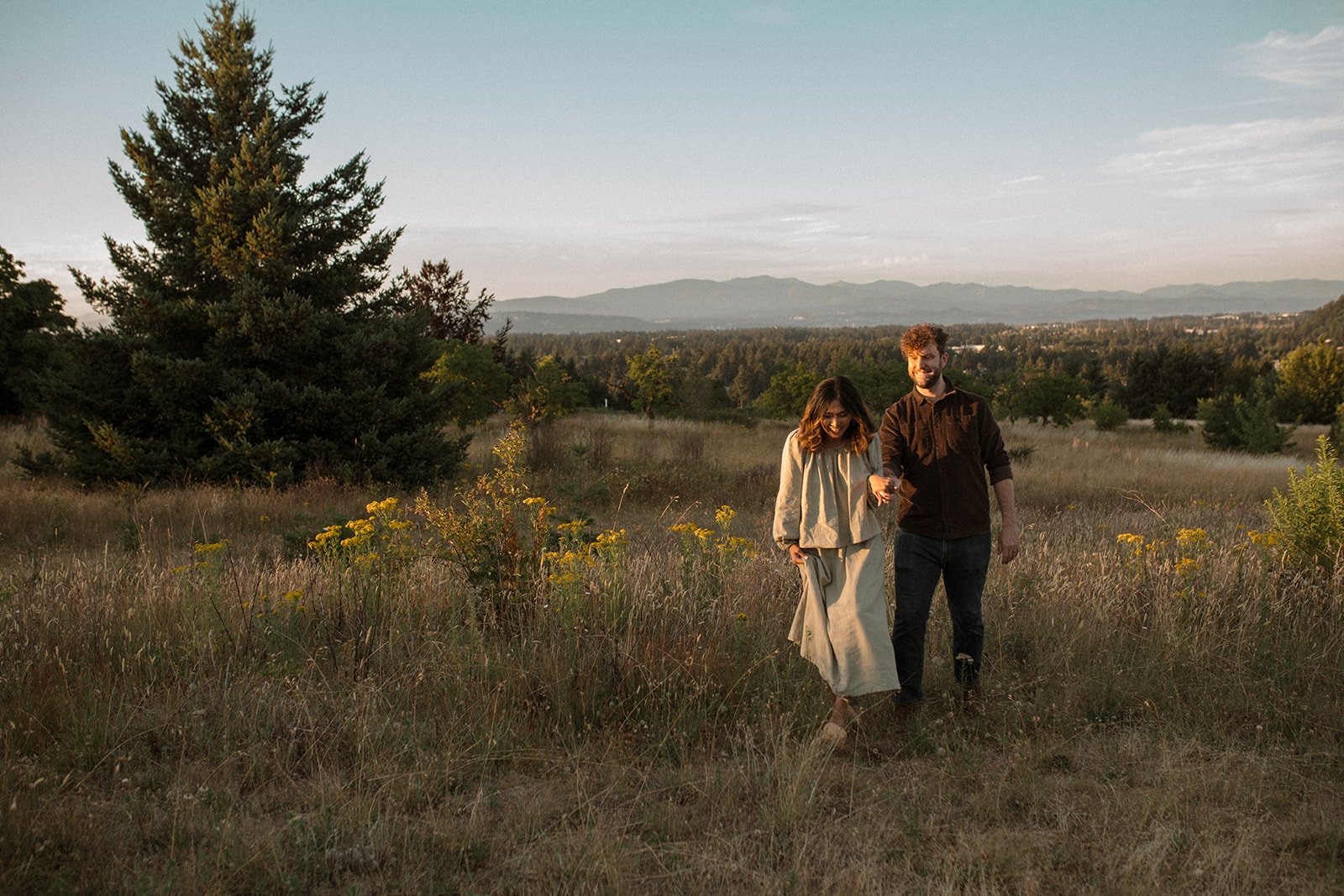 Couple walking through an open field with mountain views during a relaxed Oregon engagement session