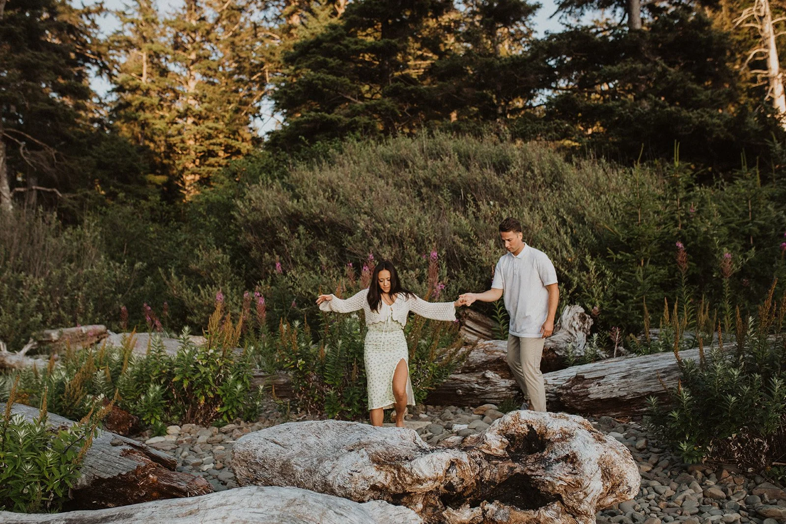 Couple walking hand in hand through driftwood and greenery during an Oregon coast engagement session