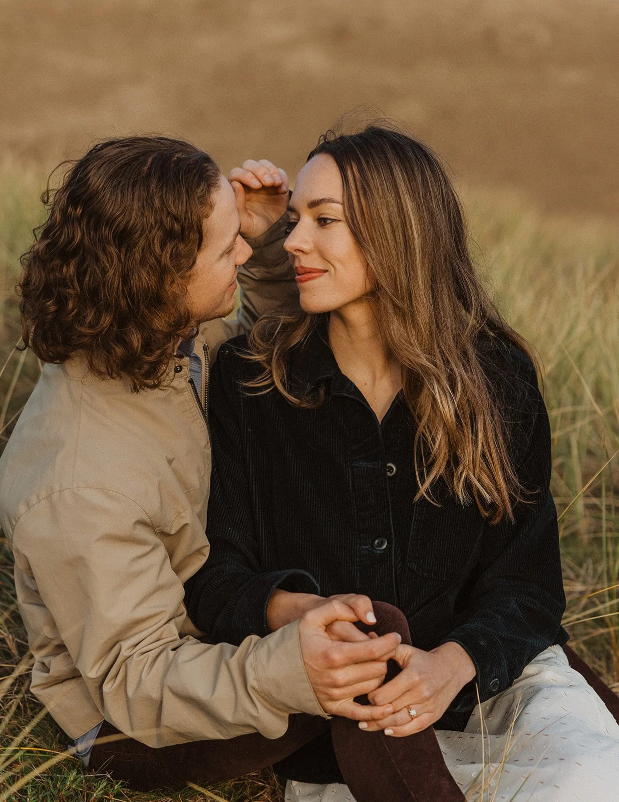 Close-up of couple sitting in tall grass at golden hour, holding hands and smiling during engagement session