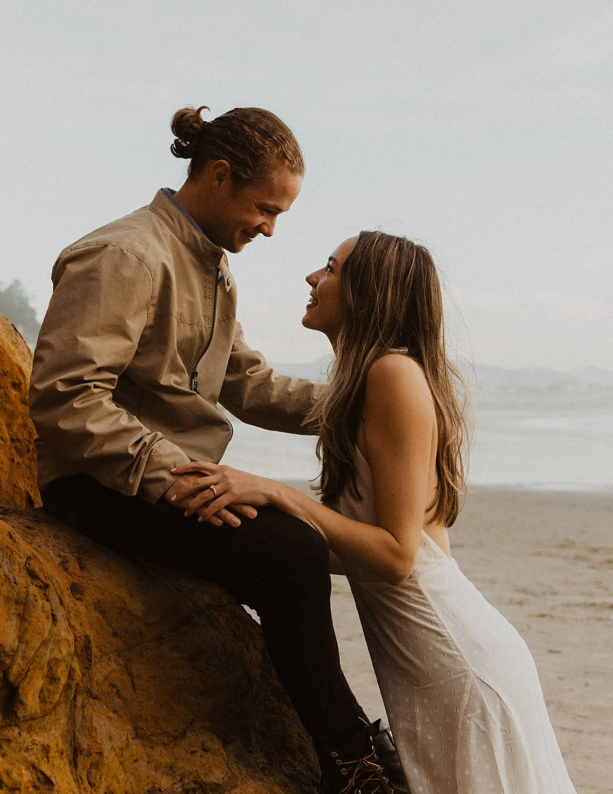 Couple sitting together on coastal rocks, smiling at each other during an Oregon Coast engagement session