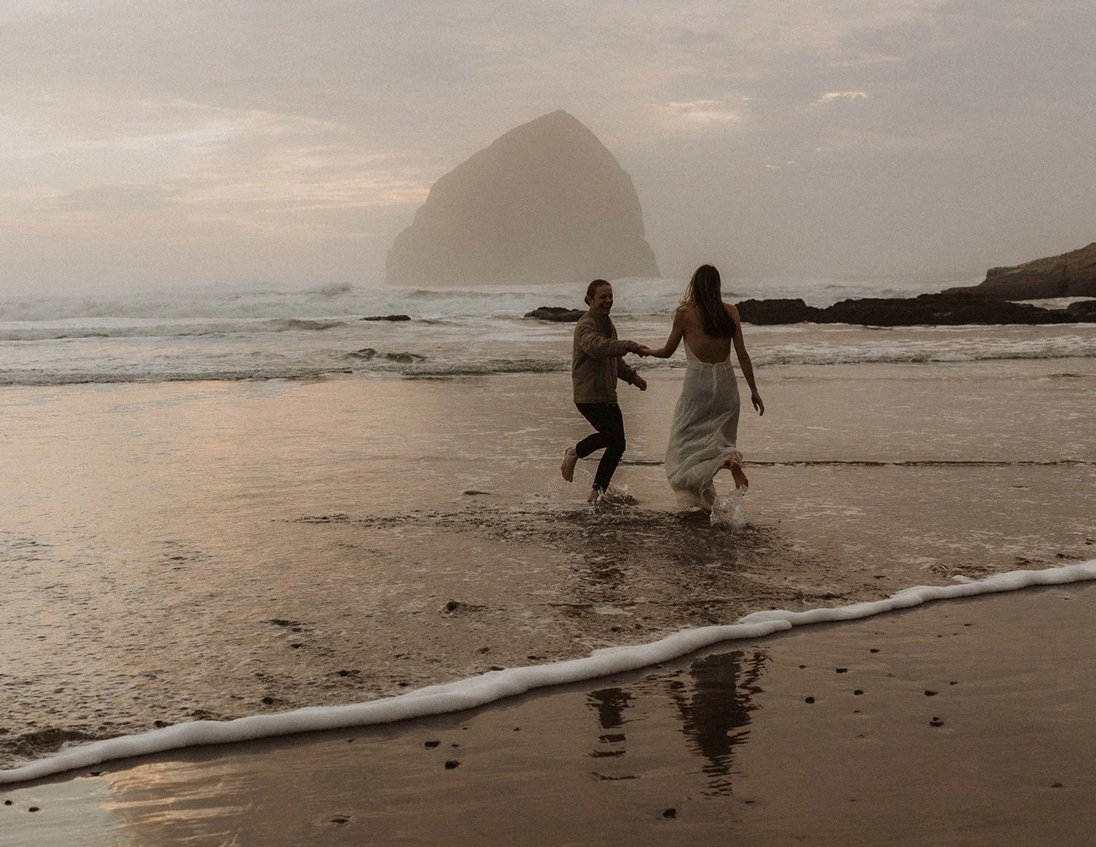 Couple running into the ocean waves near Haystack Rock during a playful Oregon coast engagement session