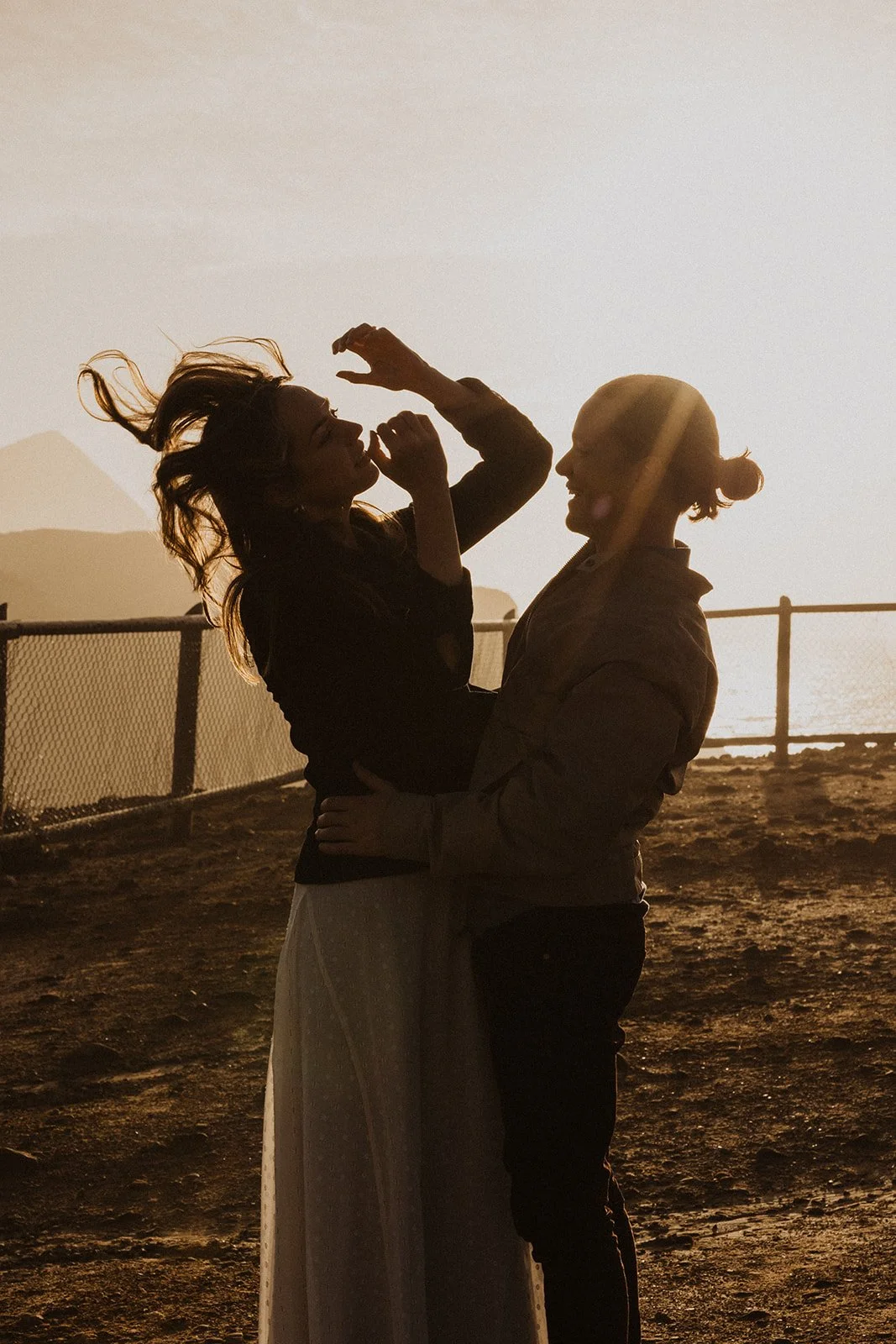 Silhouette of a couple laughing together in the wind during a golden hour engagement session on the Oregon coast