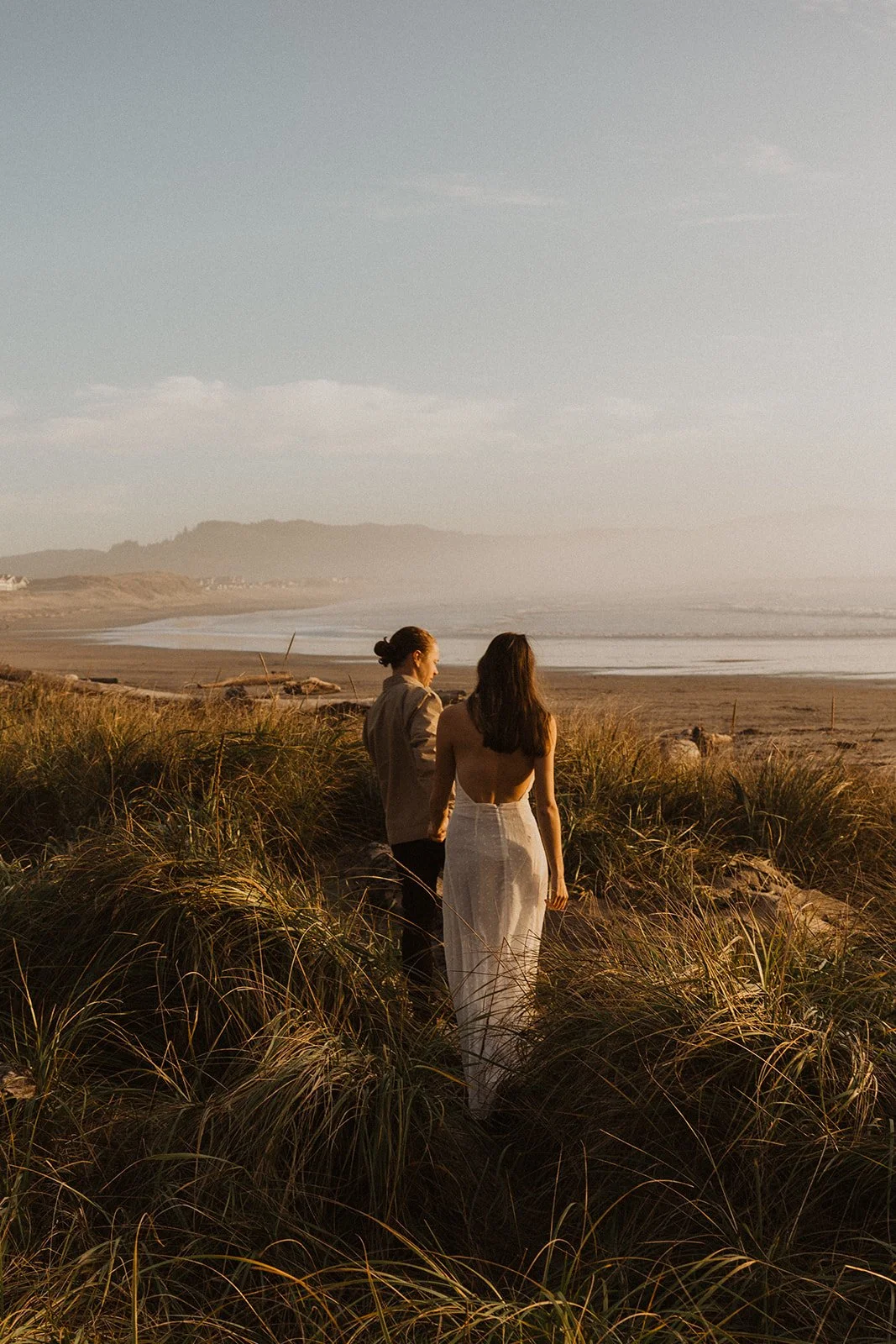 Couple walking through coastal grass dunes overlooking the ocean during an Oregon coast engagement session at sunset