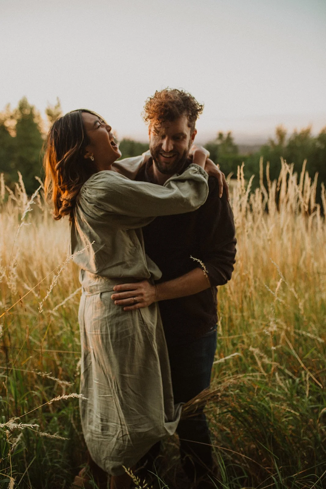 Candid couple laughing together in tall grass during golden hour meadow engagement photos with warm sunset light