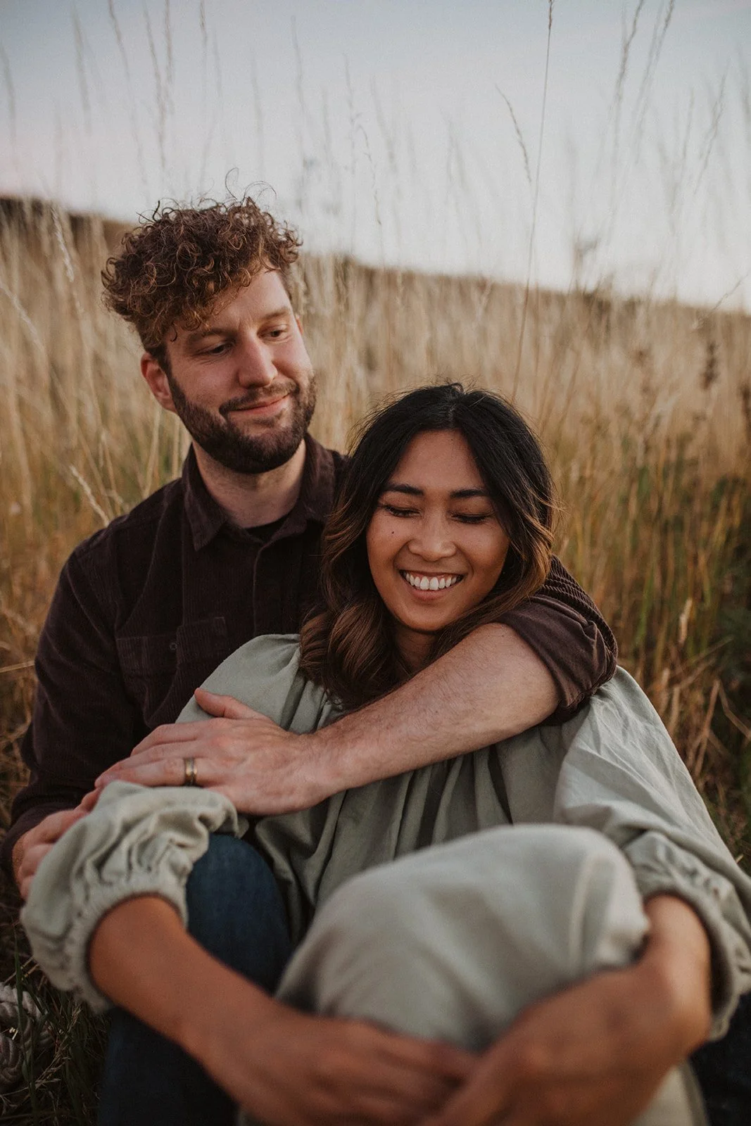 Couple sitting in tall grass embracing during golden hour engagement session in an Oregon meadow
