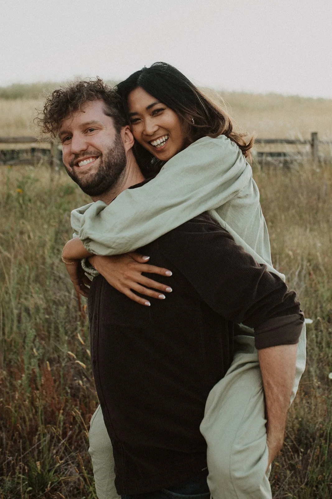 Smiling couple in a grassy Oregon meadow with woman giving piggyback ride during candid engagement session