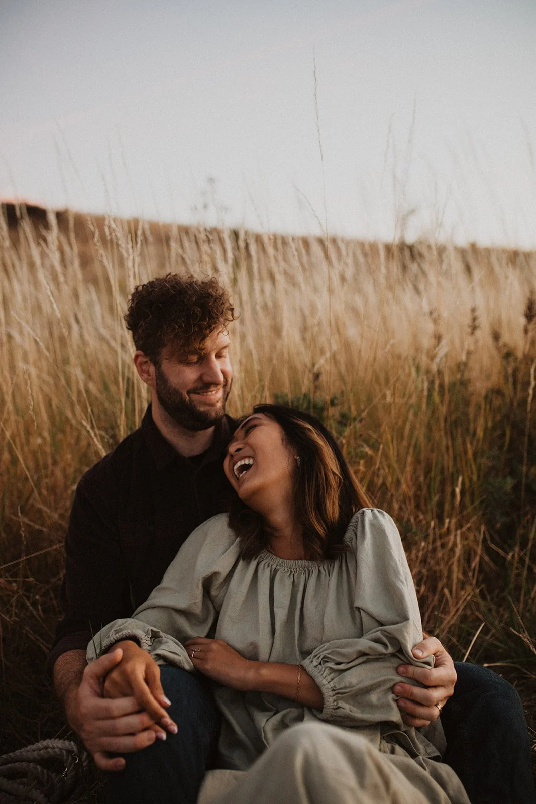Couple laughing together while seated in a meadow at sunset during a warm, candid engagement session