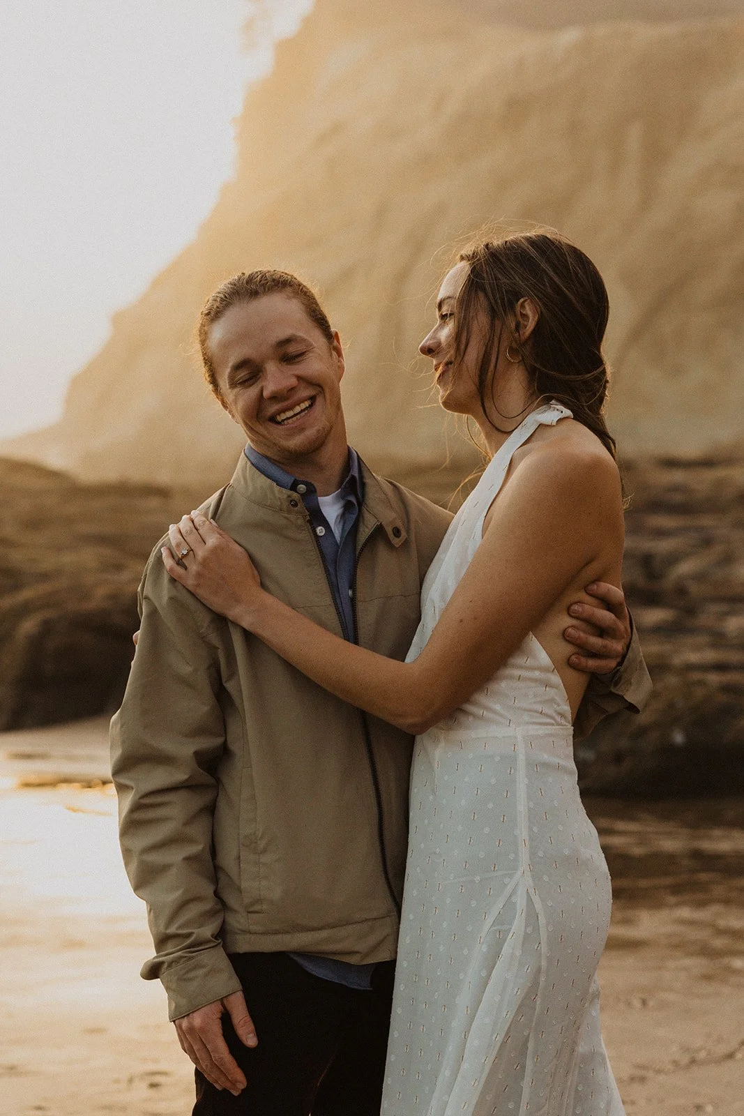 Couple embracing and smiling together during golden hour engagement session along an Oregon coast cliff