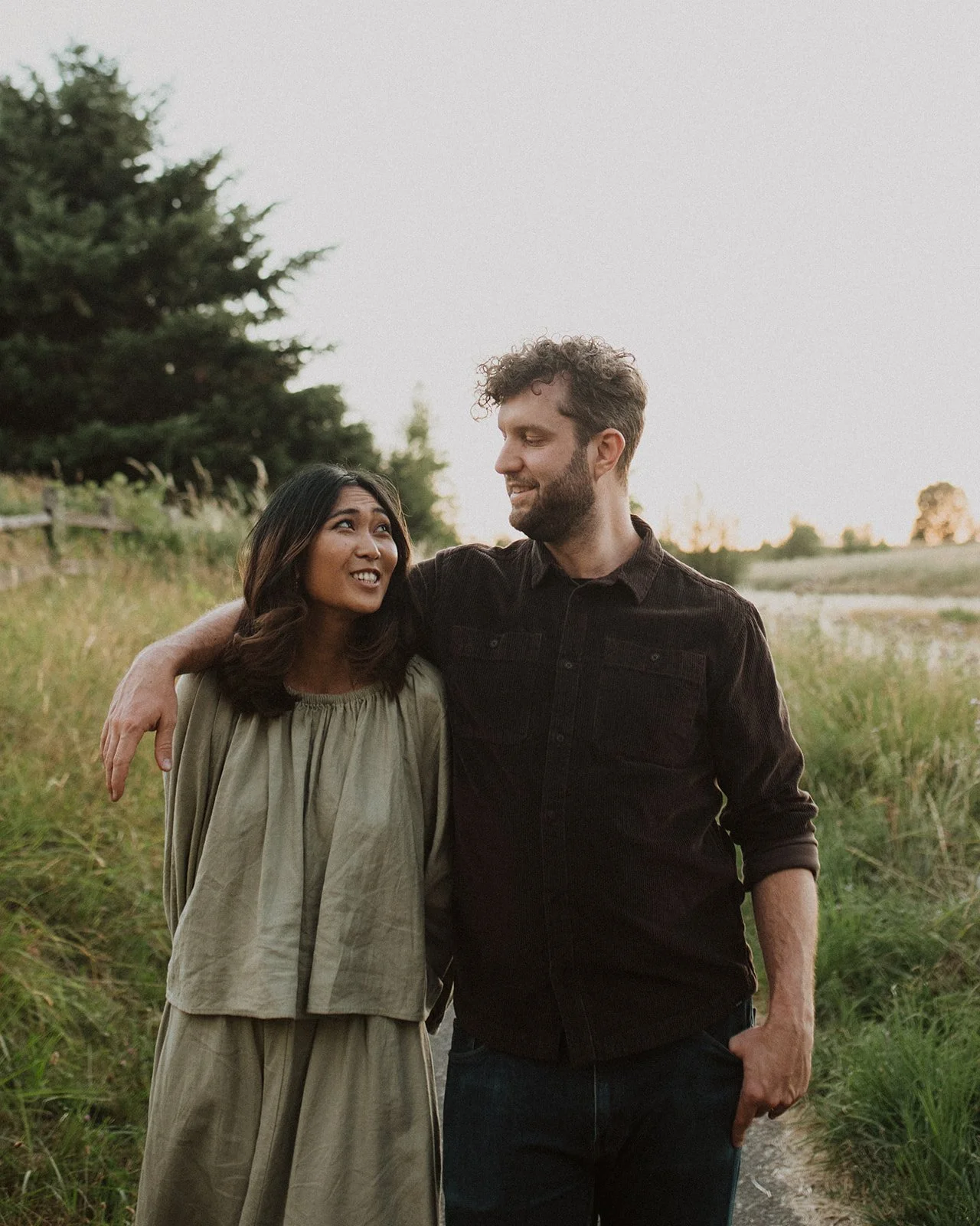 Couple walking arm in arm along a grassy path during an outdoor engagement session, smiling and talking naturally