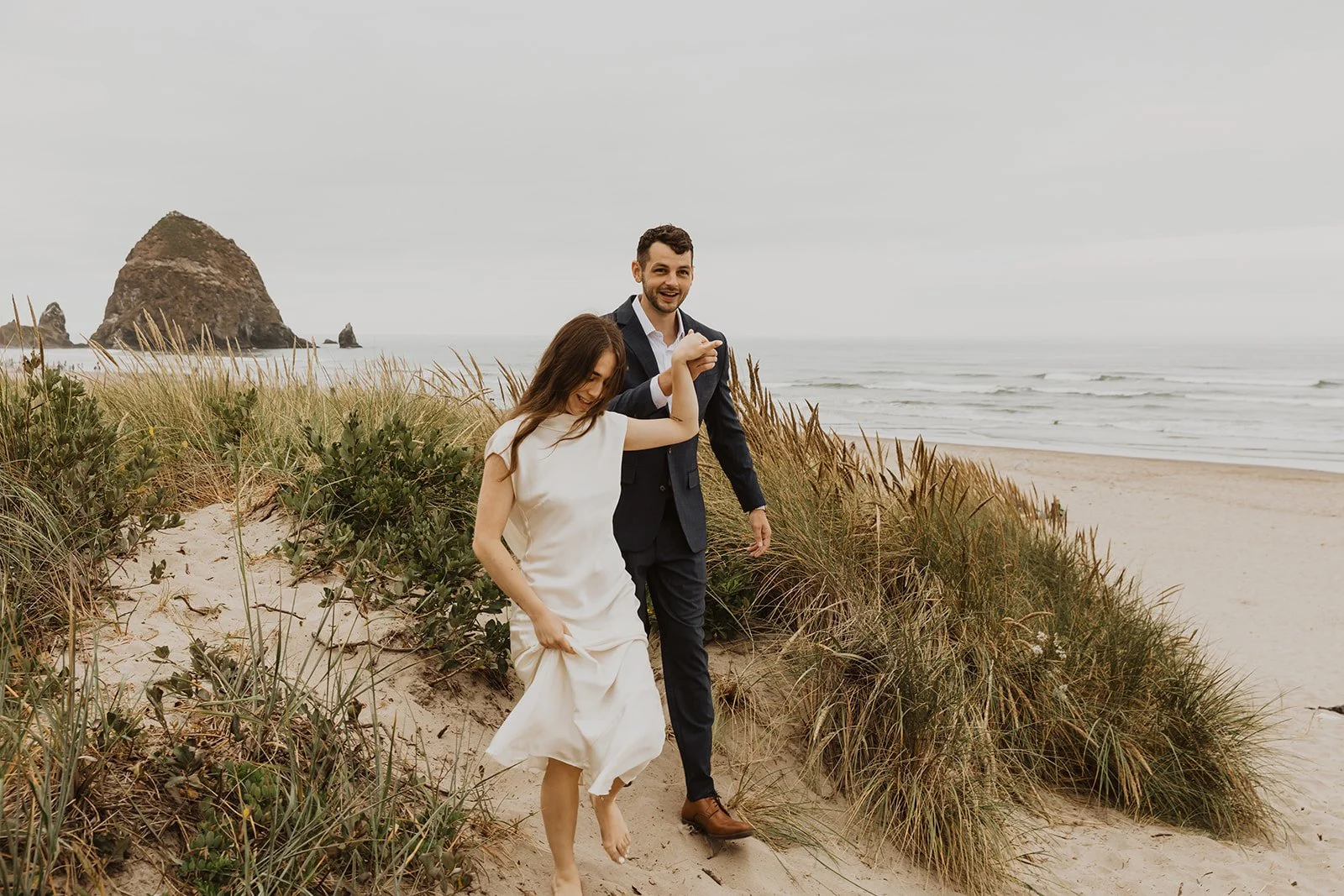 Couple walking barefoot through the sand dunes during an intimate Cannon Beach Oregon wedding with Haystack Rock in the background