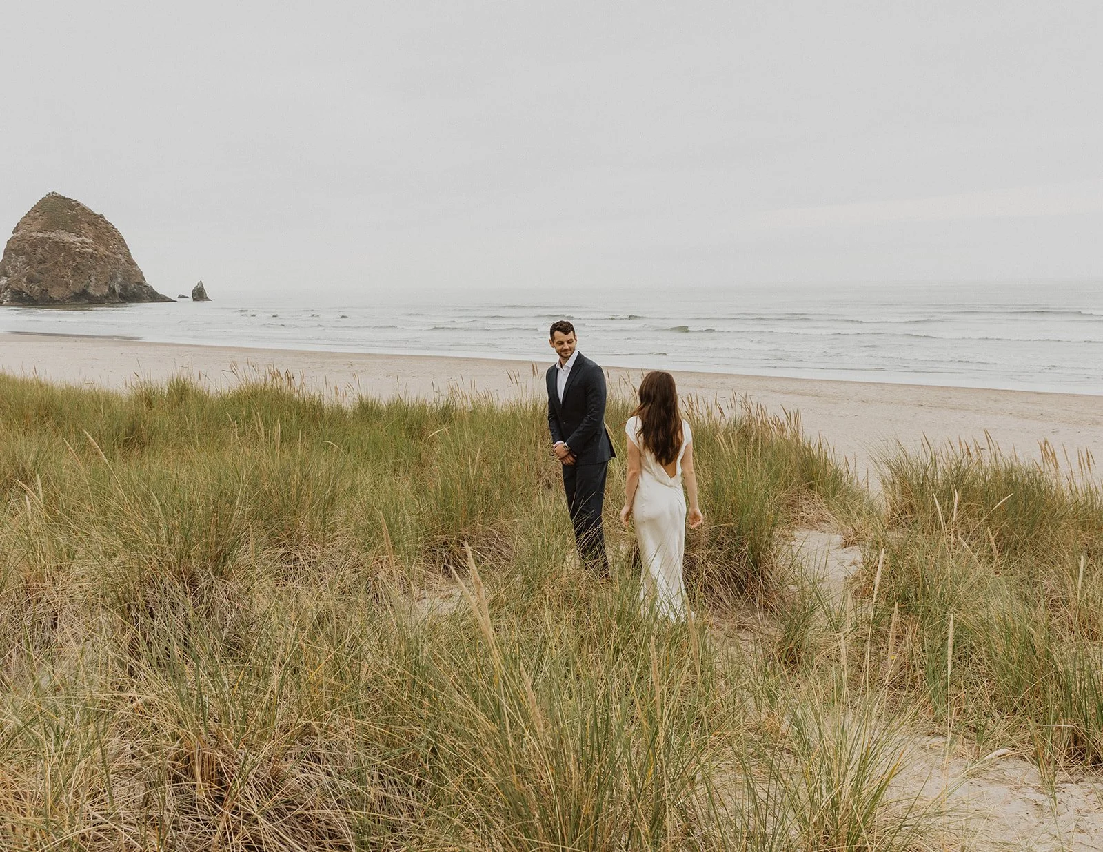 Bride and groom walking through grassy dunes during a relaxed beachfront wedding at Cannon Beach on the Oregon coast