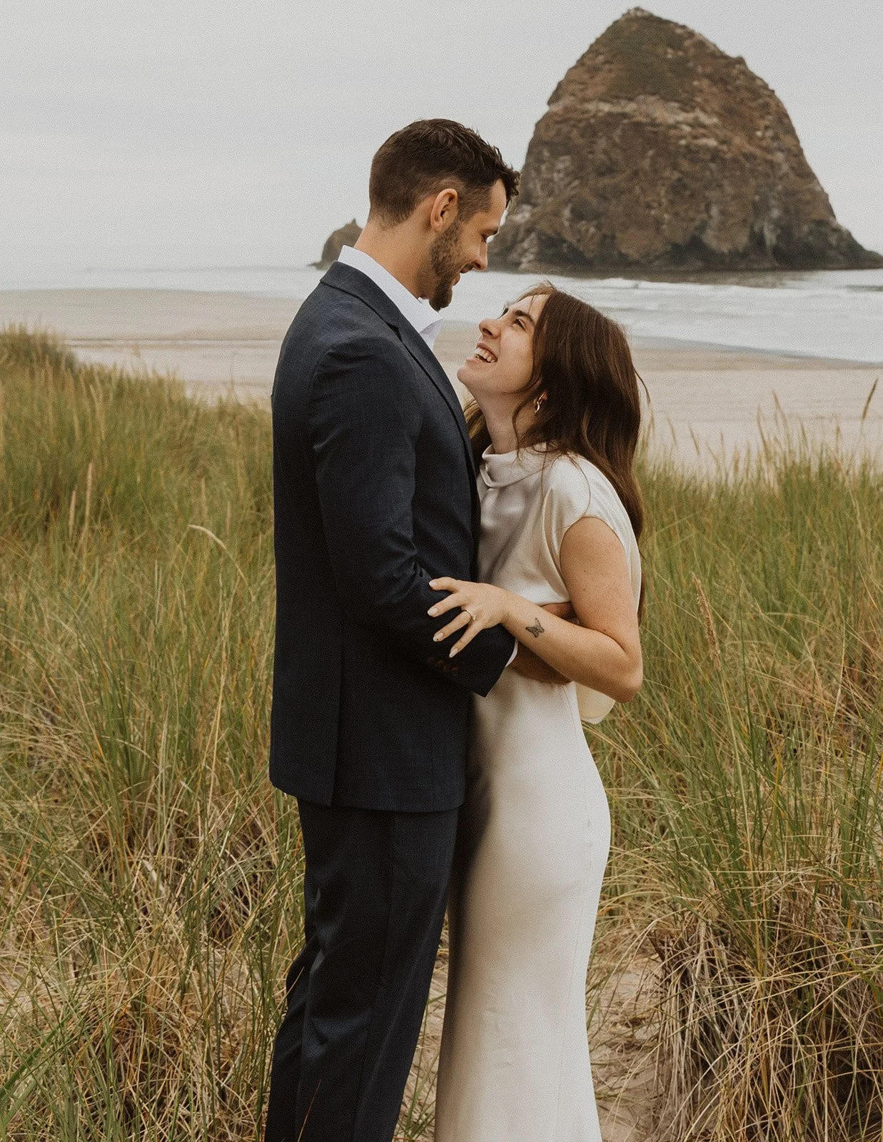 Bride and groom embracing in the beach grass with Haystack Rock in the background after their Oregon coast wedding