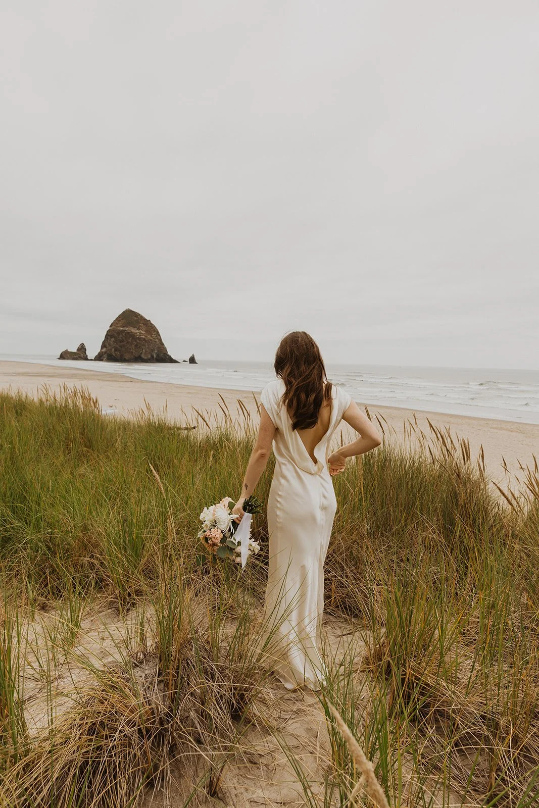 Bride walking through coastal sand dunes with her bouquet overlooking Cannon Beach and Haystack Rock