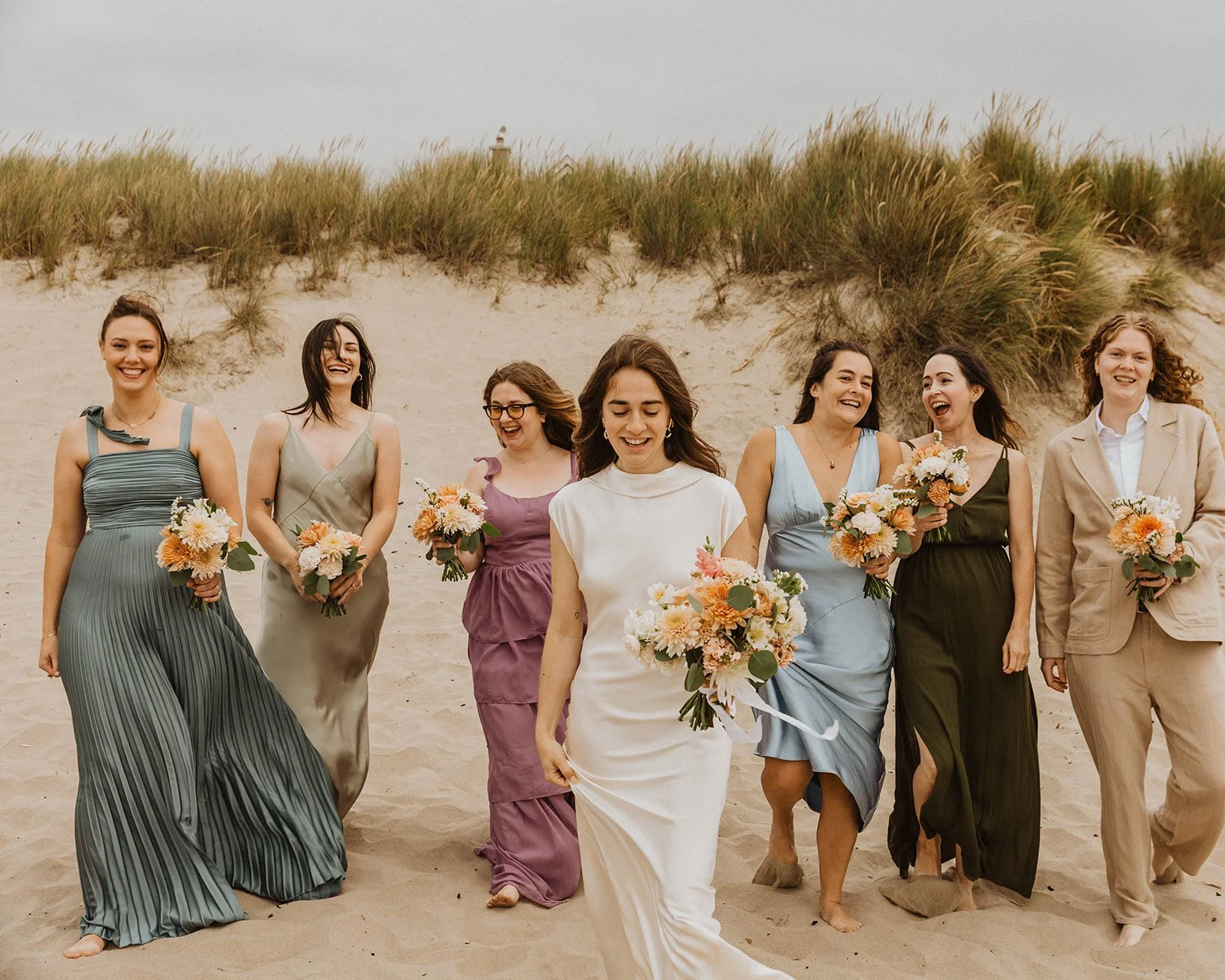 Bride and bridesmaids walking together through the sand holding pastel wedding bouquets at Cannon Beach