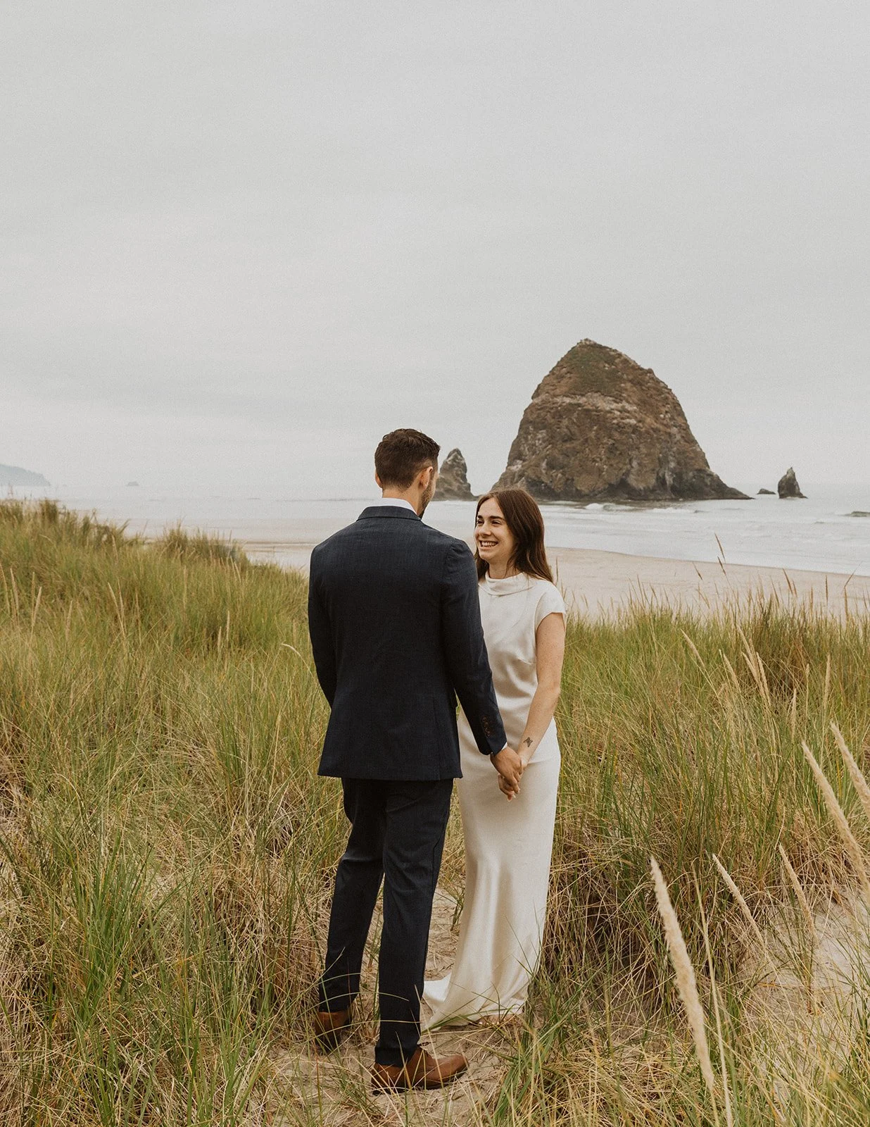 Bride and groom holding hands in the coastal dunes at Cannon Beach with Haystack Rock in the background