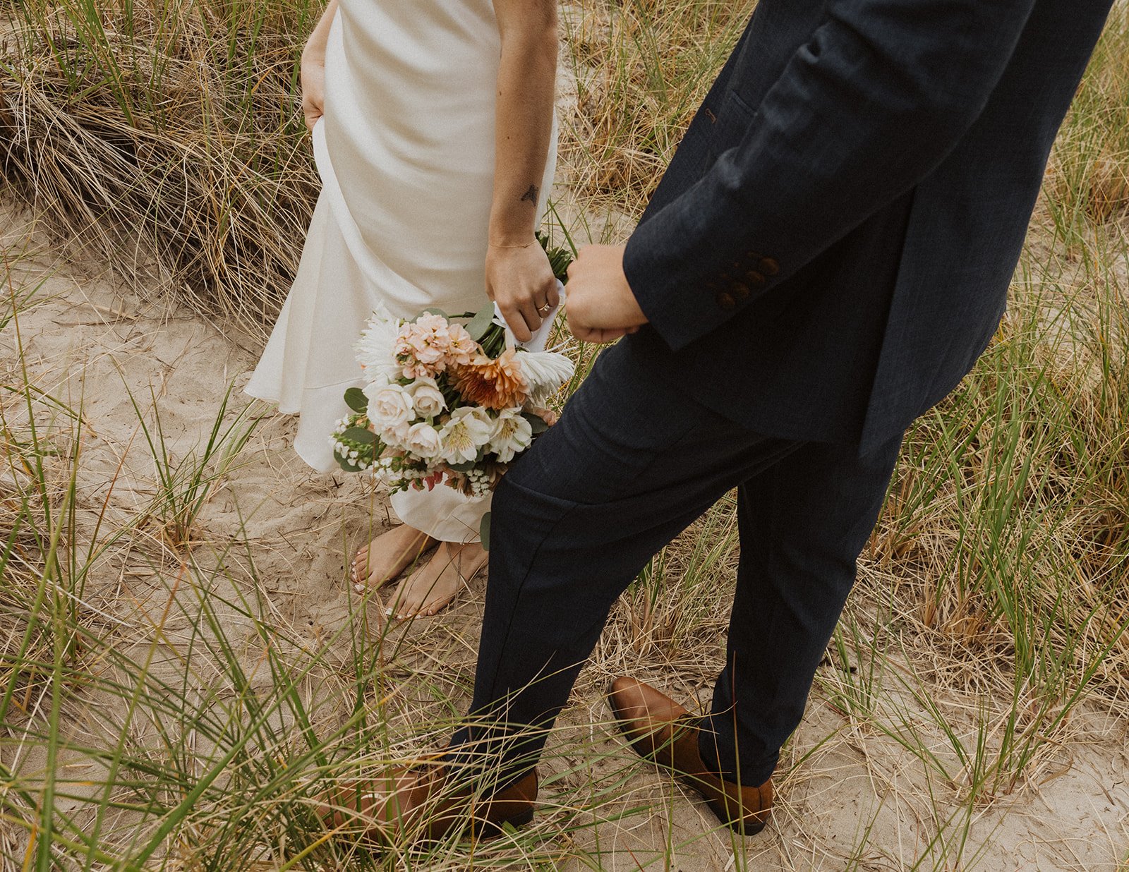 Close-up detail of a bride and groom walking barefoot through sandy coastal dunes with an organic wedding bouquet