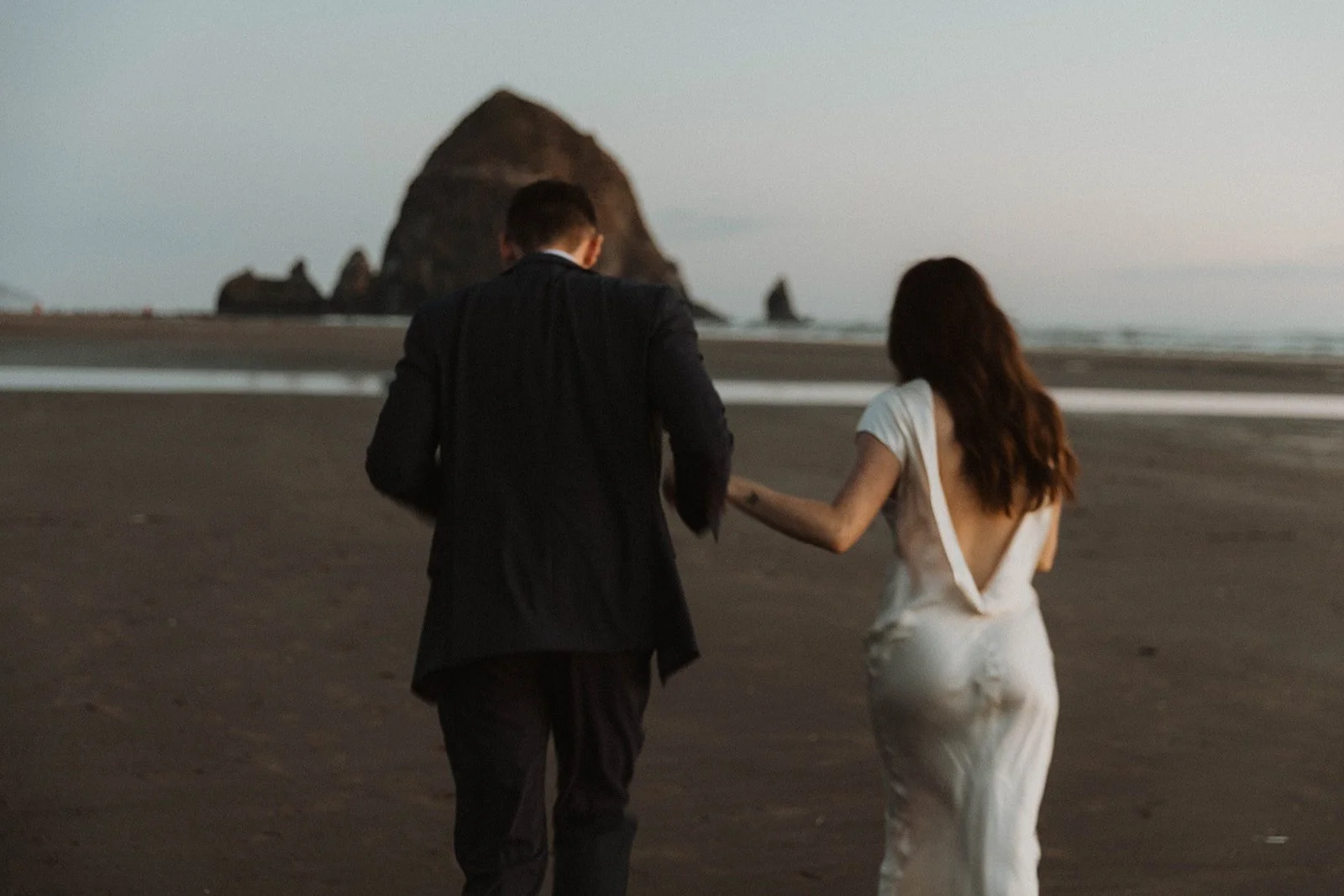 Couple walking hand-in-hand across Cannon Beach at dusk after their intimate elopement