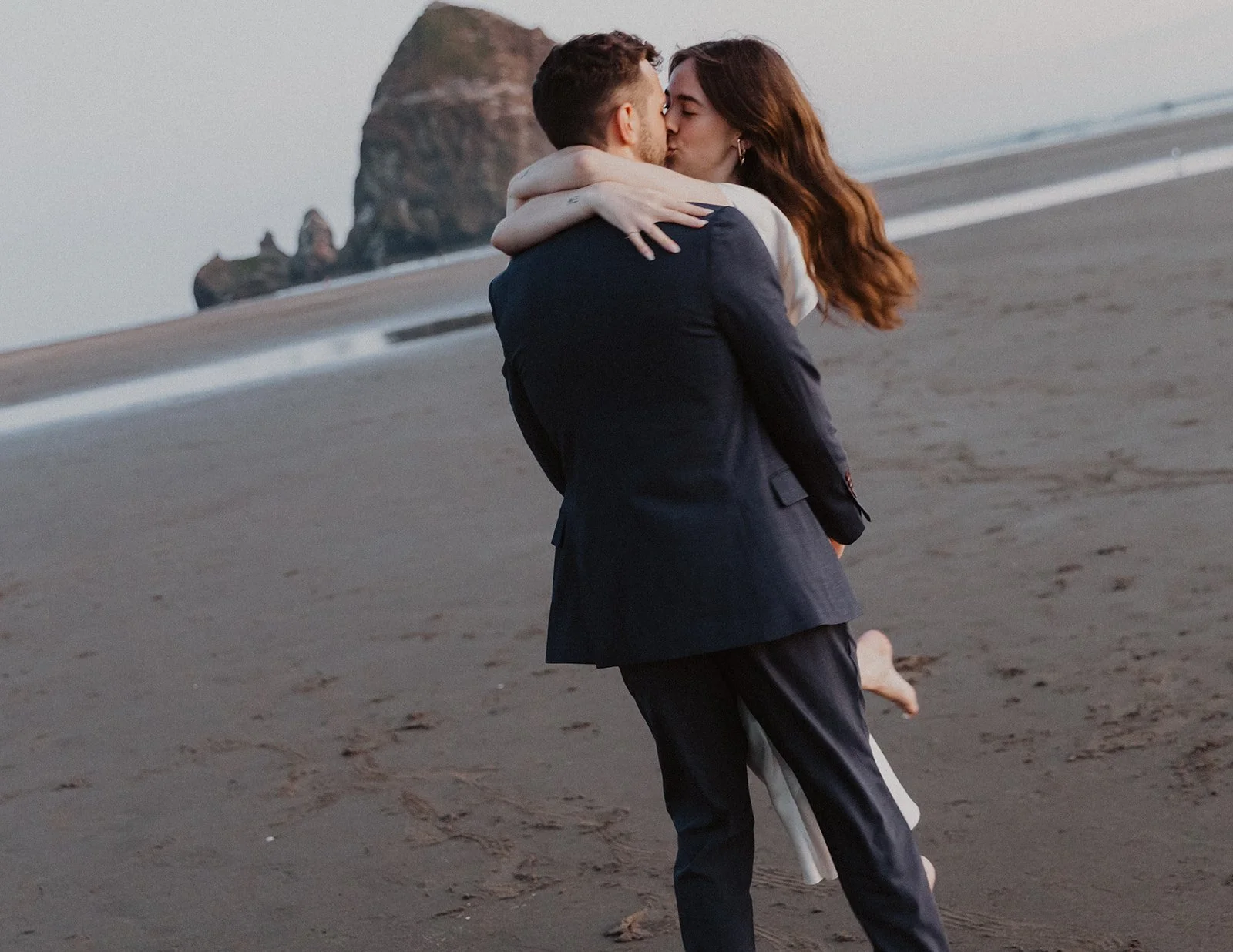 Bride wrapping her arms around groom during a playful kiss on Cannon Beach with Haystack Rock in the background