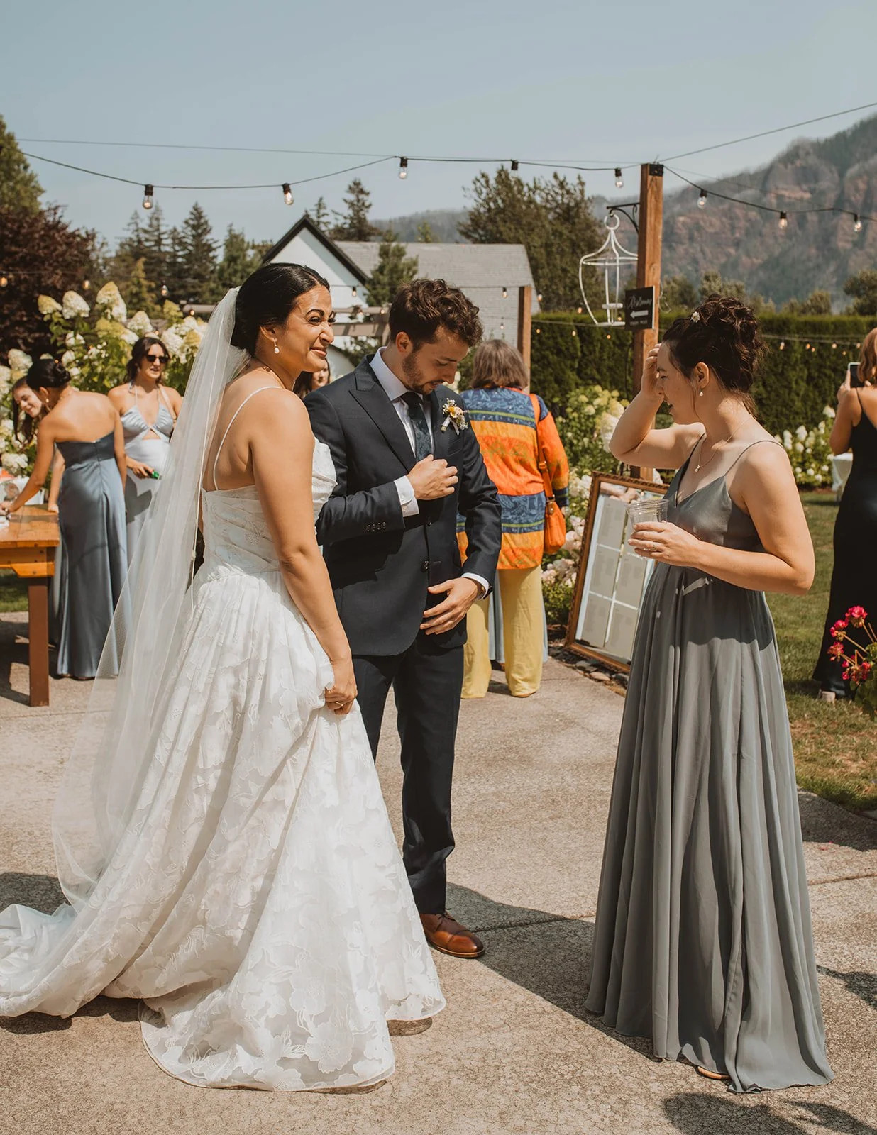Bride and groom chatting with a bridesmaid during outdoor cocktail hour at Cape Horn Estate, surrounded by string lights, mountains, and hydrangea gardens.