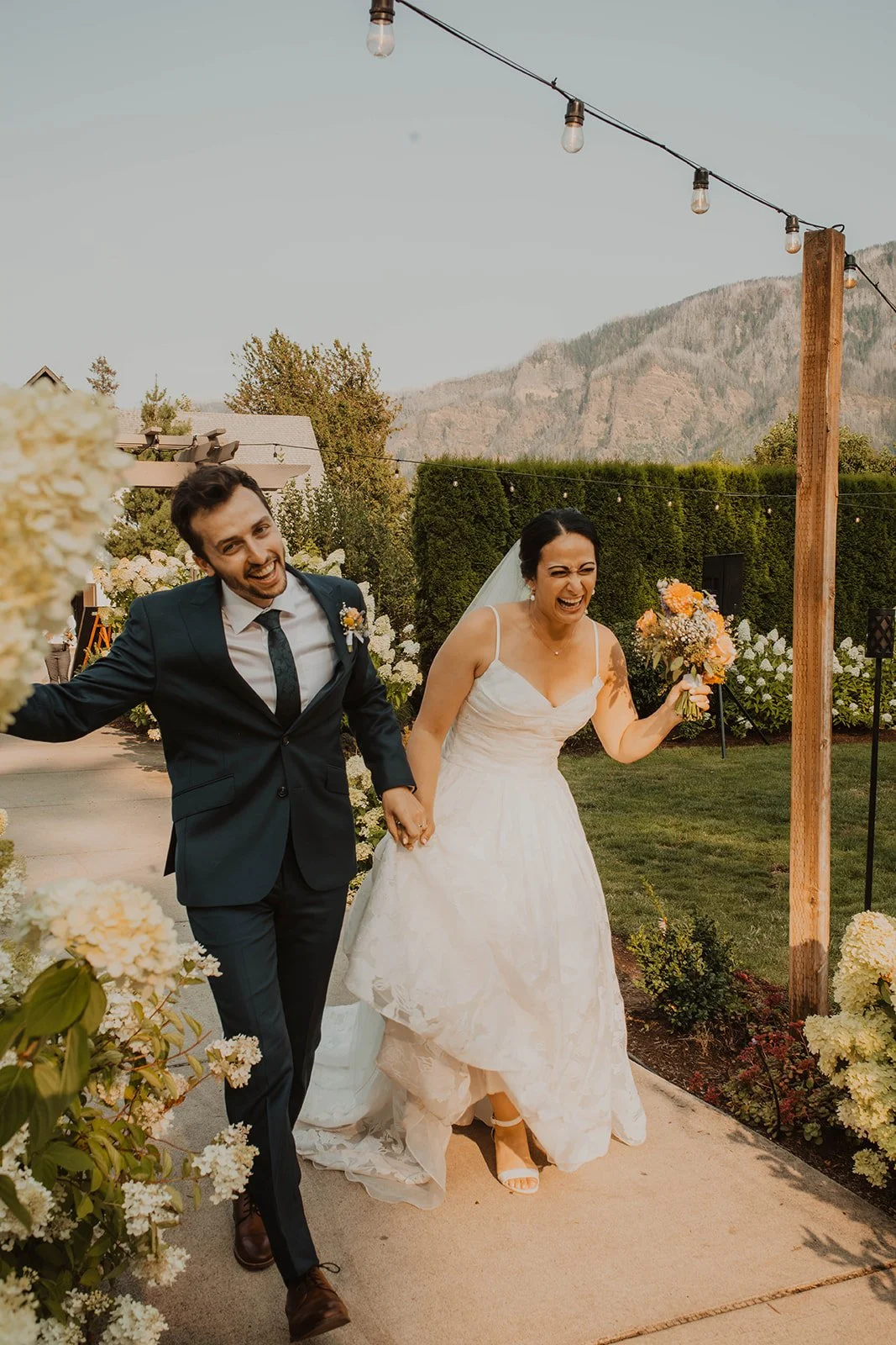 Just married couple laughing and running hand in hand through the garden pathways at Cape Horn Estate with mountain views and white hydrangeas surrounding them.