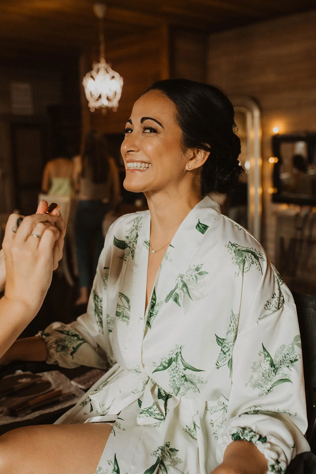 Bride smiling during wedding morning hair and makeup while wearing a white floral robe in a warm, cozy getting-ready suite.