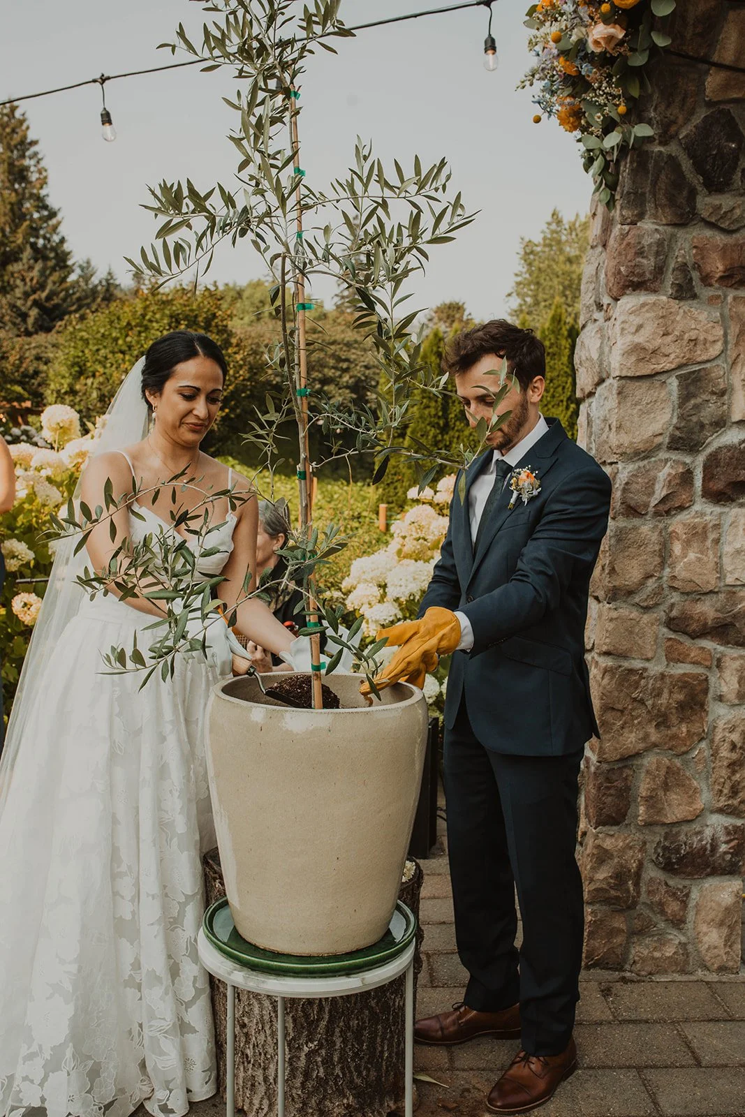 Bride and groom participate in an olive tree planting unity ceremony beside a stone arch decorated with colorful wedding flowers.