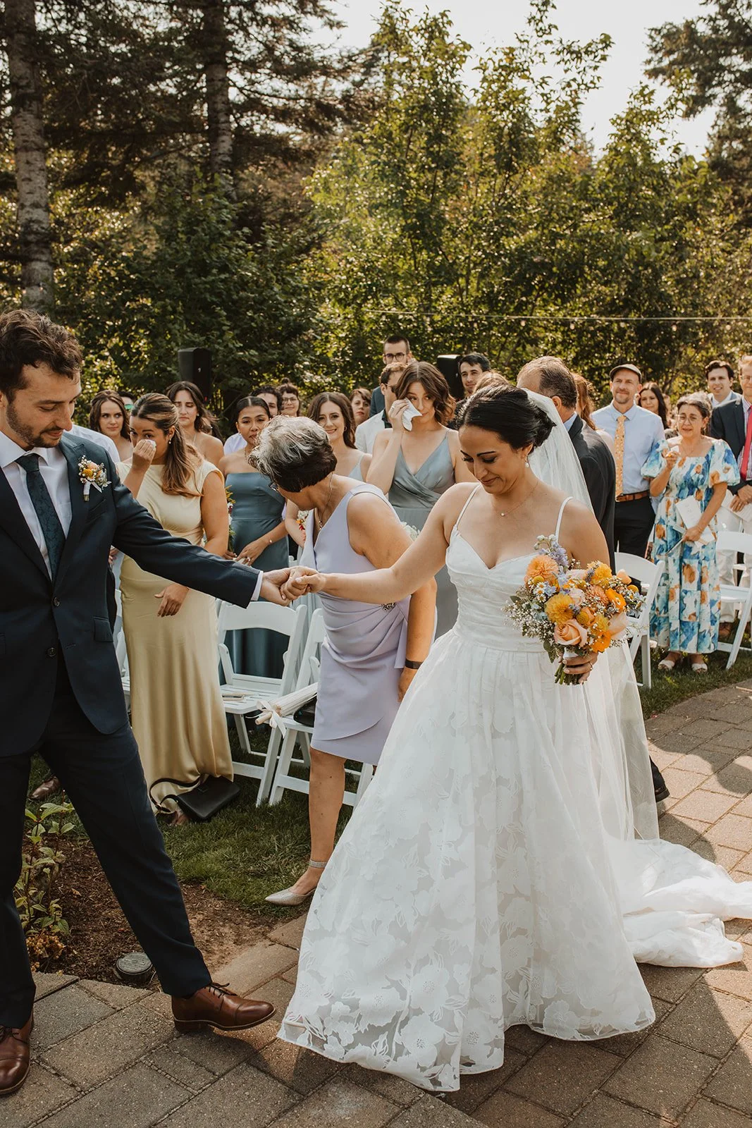 Newly married couple walk hand in hand down the aisle after their outdoor ceremony while emotional guests smile and wipe away tears in the background.